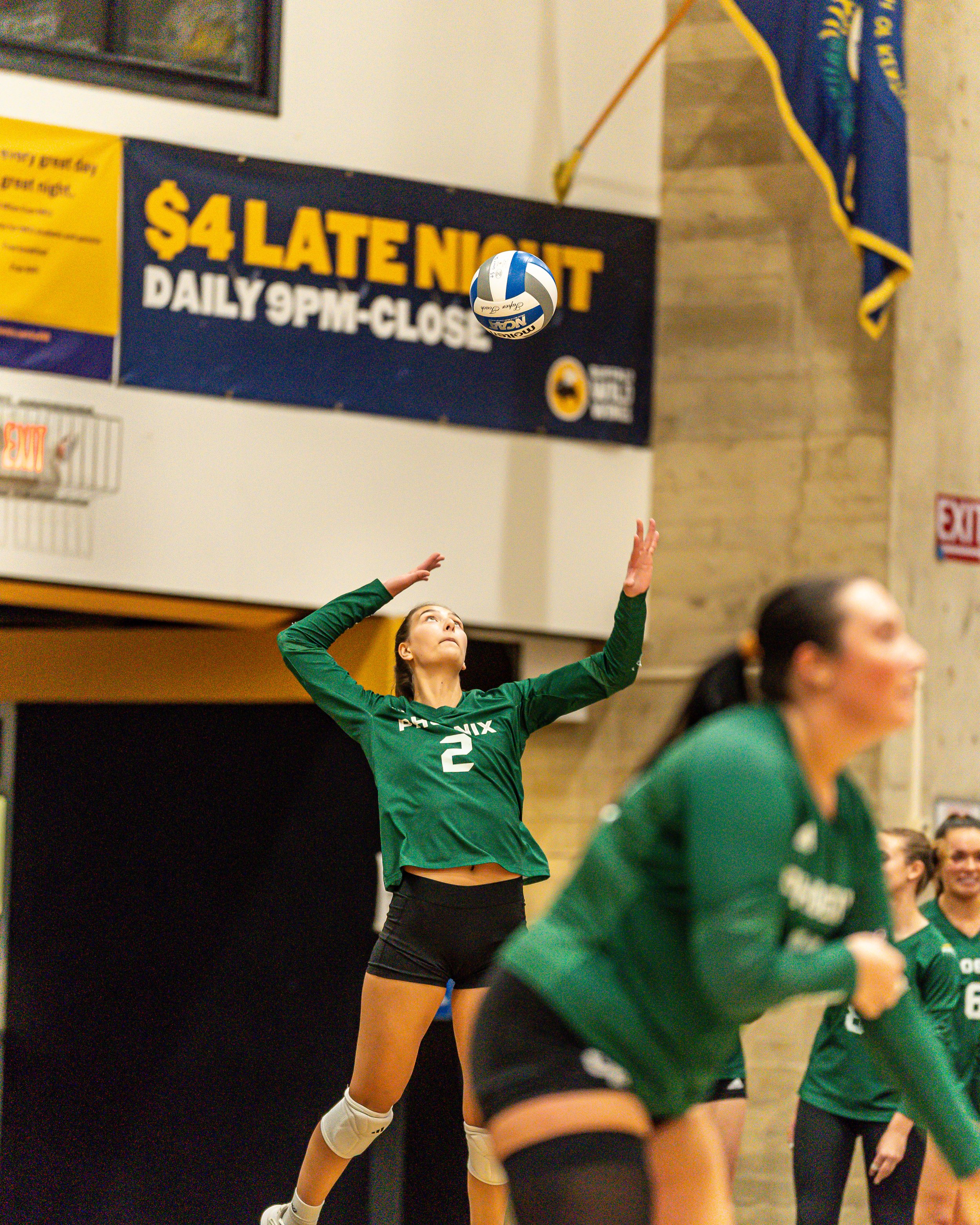 A female volleyball player in a green uniform, number 2, jumping to hit the ball during a game in an indoor gym.