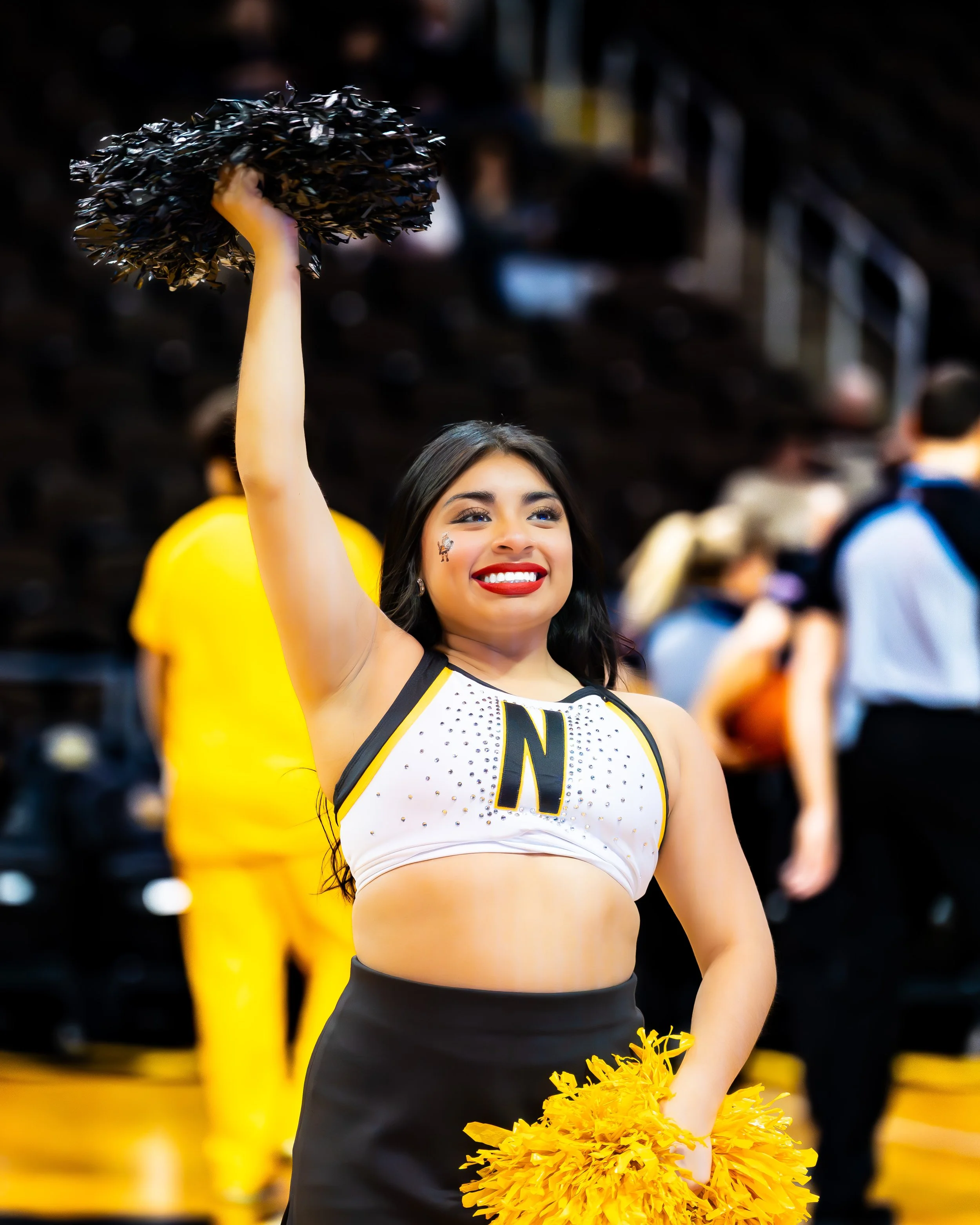 A cheerleader in a white and black uniform with a large letter 'N' on her top, holding black pom-poms in her right hand and yellow pom-poms in her left hand, smiling and looking cheerful at a sports event.