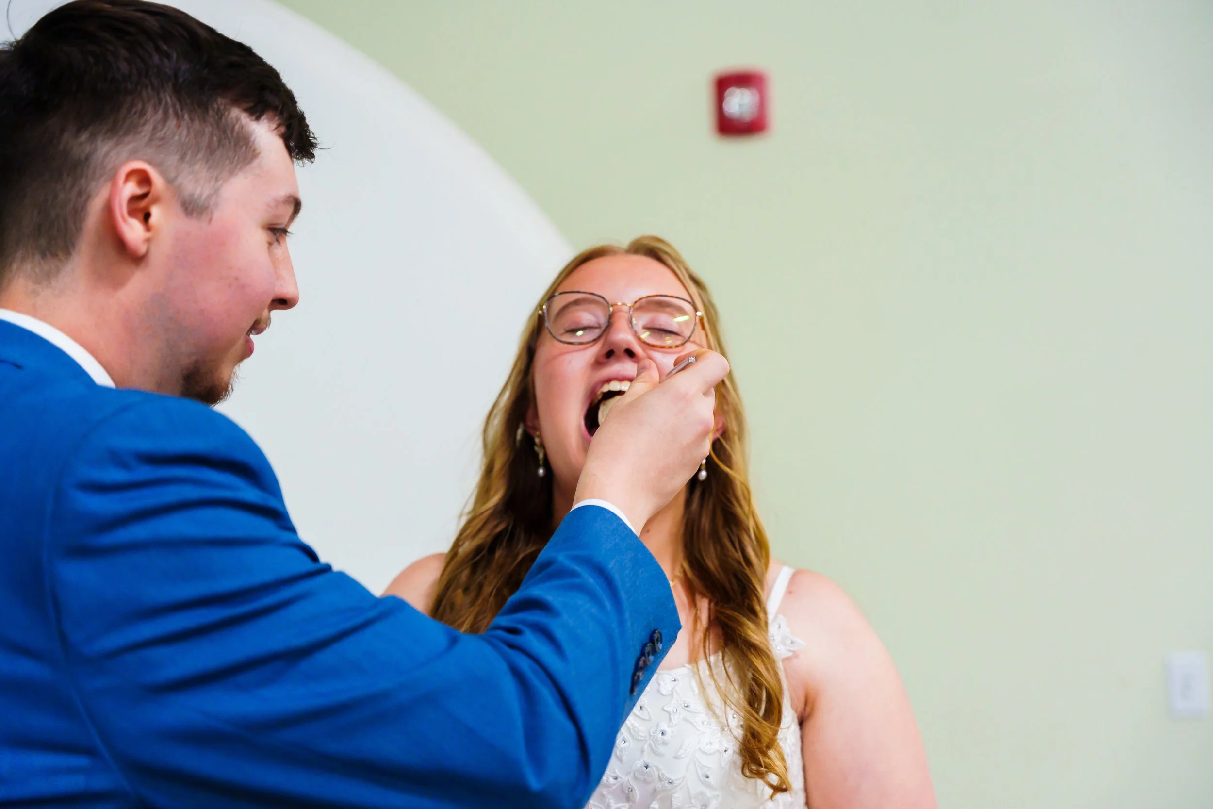 A young man with dark hair, wearing a blue suit, is smiling as he helps a young woman with red hair and glasses, who looks happy and is wearing a white dress. They are in a room with light green walls.