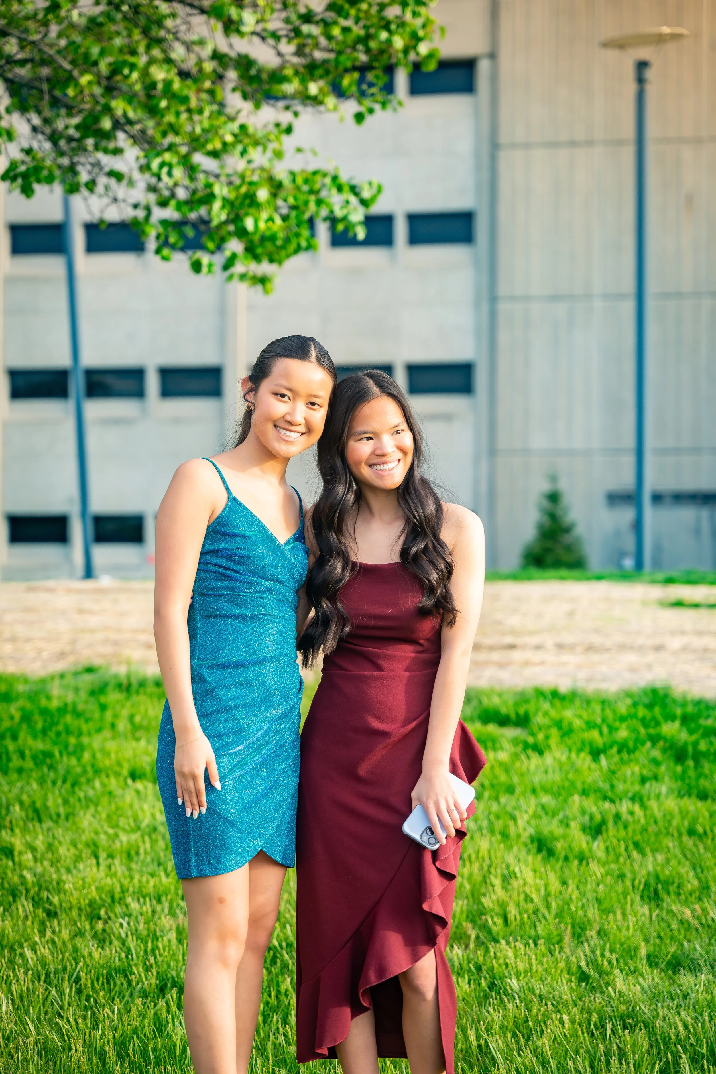 Two young women in formal dresses standing outdoors on a well-manicured lawn, smiling at the camera with a modern building and a tree in the background.