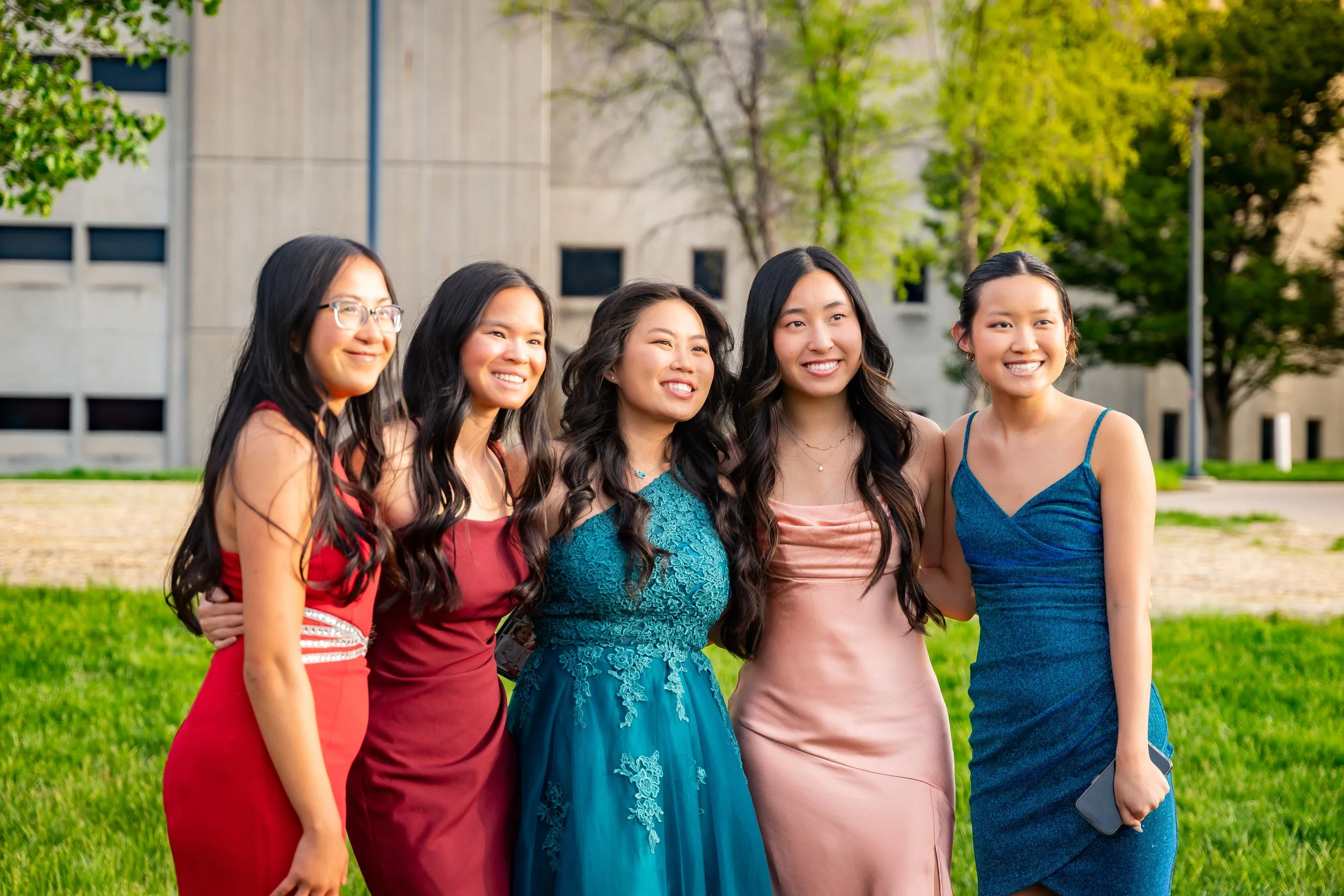 Five young women in formal dresses standing outdoors on a sunny day, smiling, with trees and a building in the background.