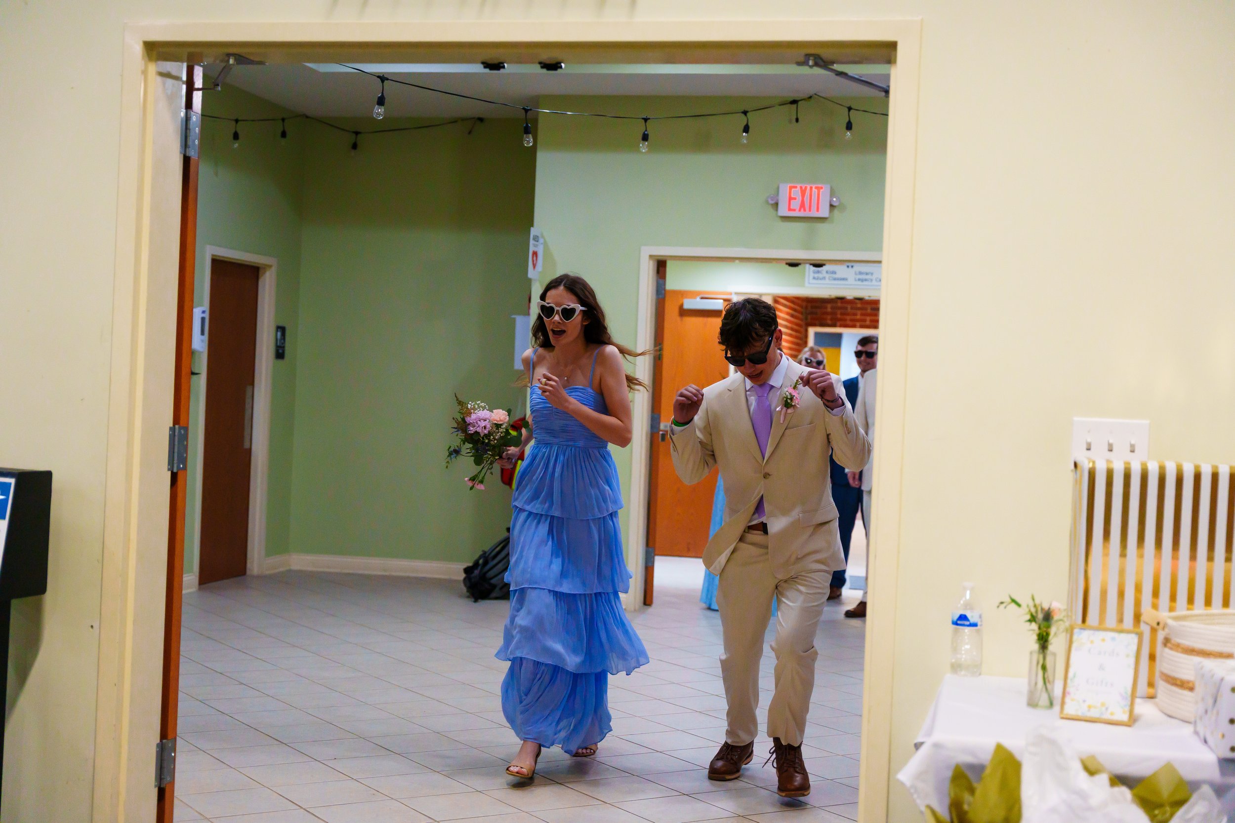 A woman in a blue dress and a man in a beige suit walk through an indoor space, with the woman holding a bouquet of flowers and wearing heart-shaped sunglasses. Several people are visible in the background, some wearing sunglasses. The setting appear