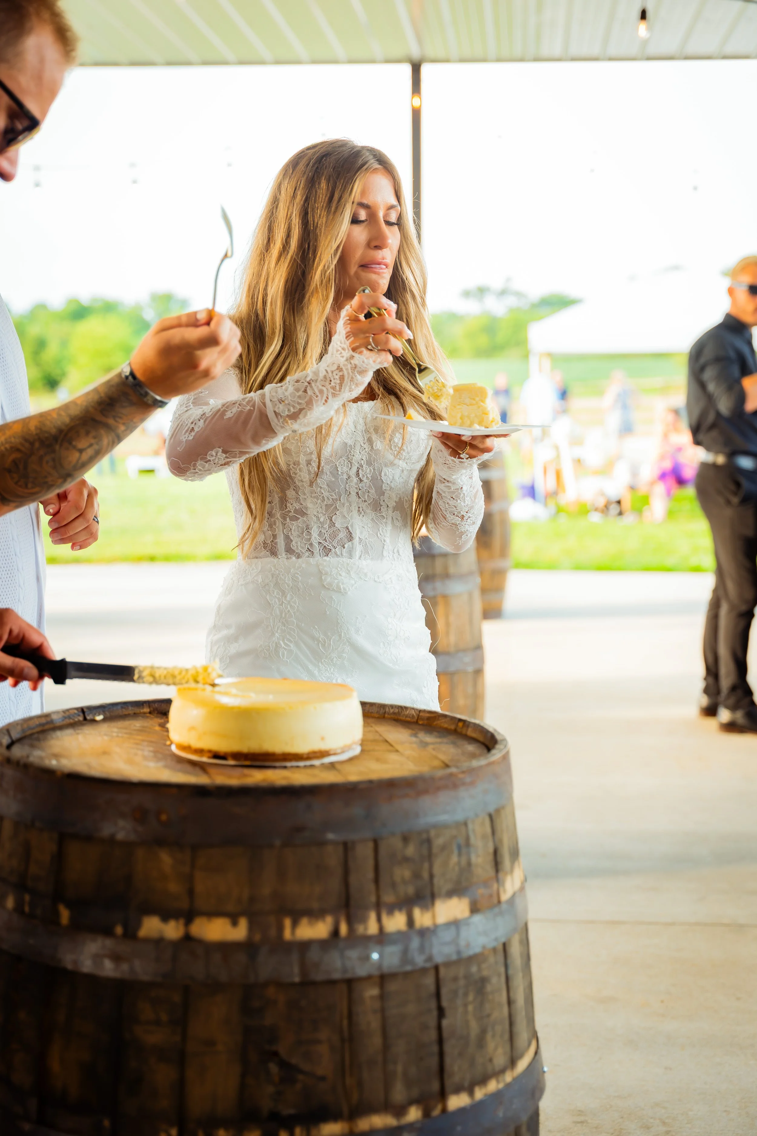 Woman in a white lace dress eating a slice of cake at an outdoor event, with a cake on a wooden barrel in front of her and people in the background.