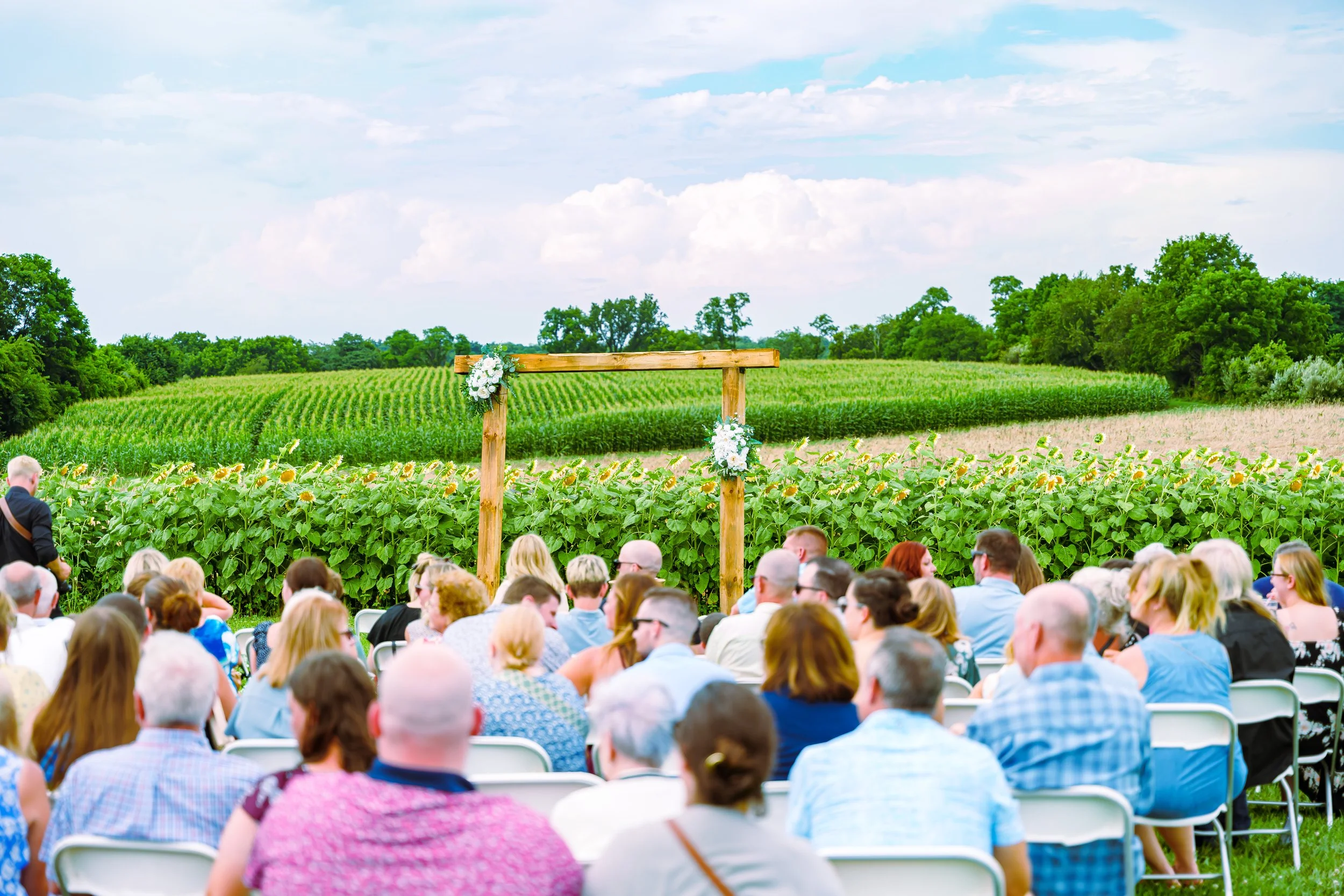 Crowd attending an outdoor event on a sunny day with green fields and trees in the background, and a floral decorated wooden arch in the distance.