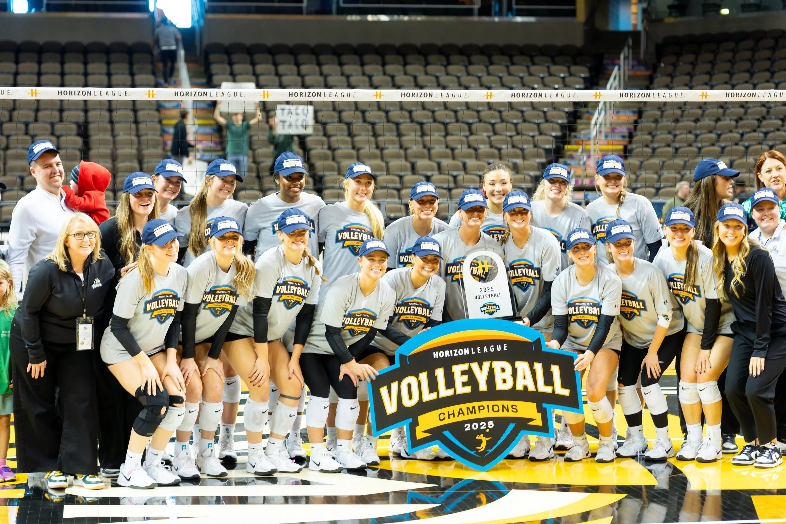 A volleyball team celebrating their championship win in 2025, holding a large sign that reads 'Horizon League Volleyball Champions 2025', with players and coaches standing on a volleyball court.