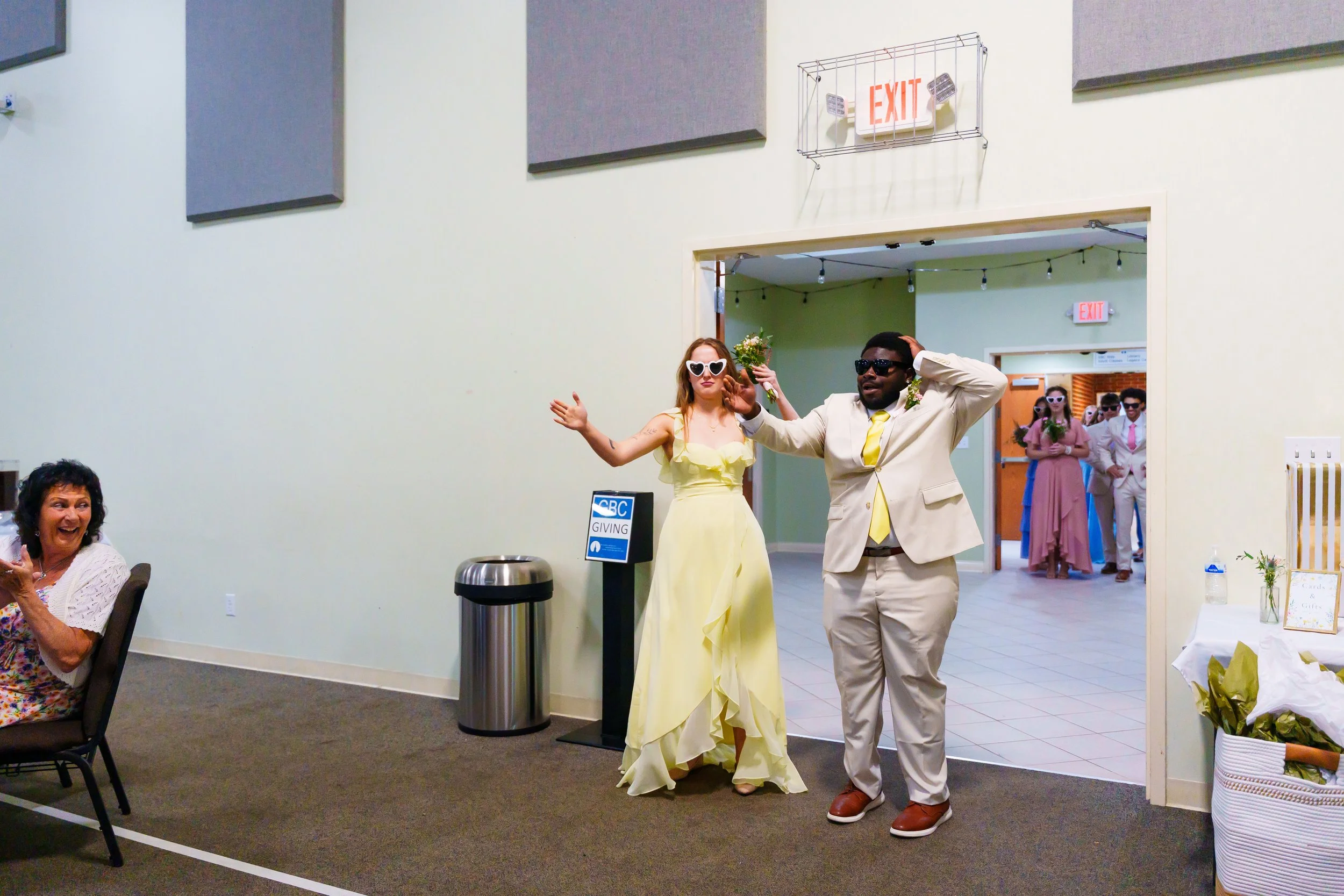 Two people stand in front of a wedding reception, dressed in formal attire; the woman wears a yellow gown and heart-shaped sunglasses, the man wears a beige suit and dark sunglasses, both wearing flower garlands. In the background, there are more gue
