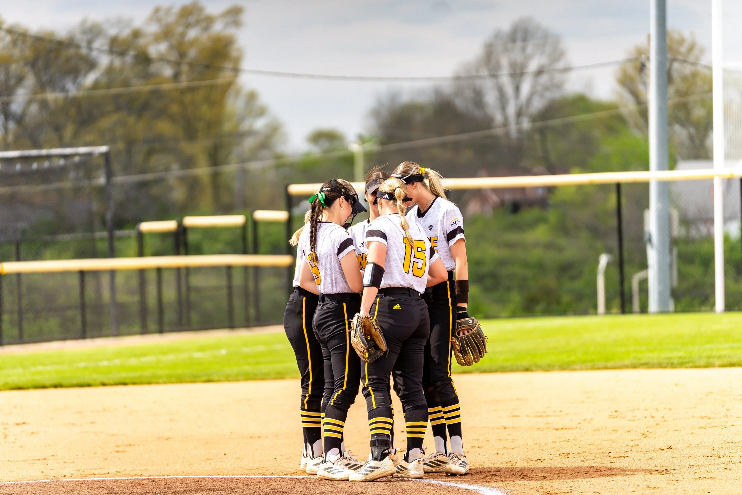 A group of female softball players huddled together on the field during a game, wearing matching uniforms with black and yellow accents.