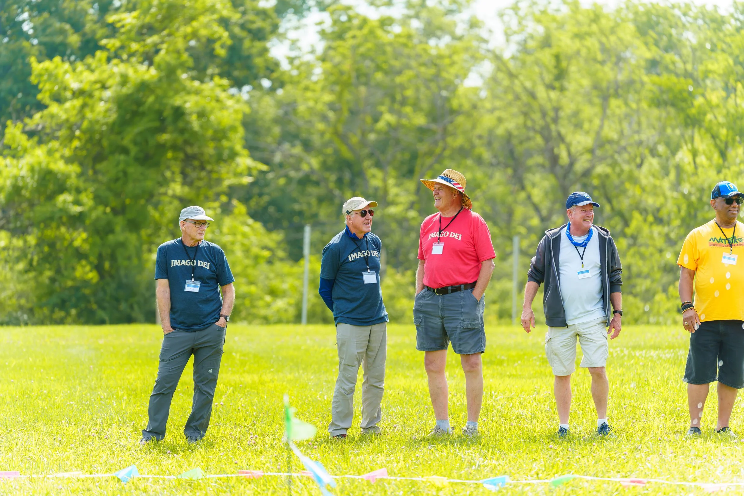 Group of five older men standing on a lush green grass field, engaging in conversation, with trees in the background. Some are wearing hats, sunglasses, and casual outdoor clothing.