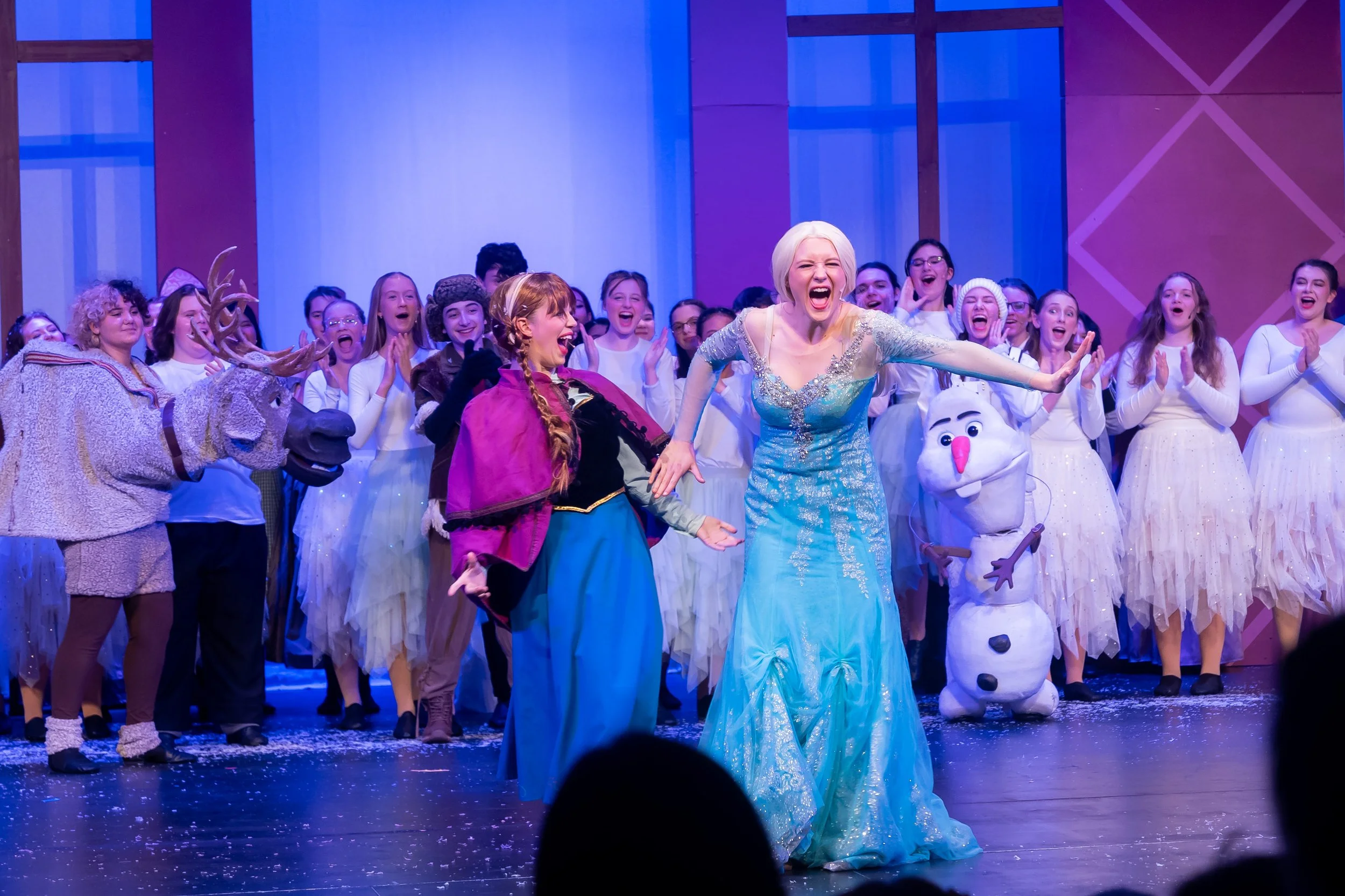Scene from a theatrical stage with performers dressed as characters from Frozen, including Elsa and Olaf, surrounded by a group of smiling cast members.