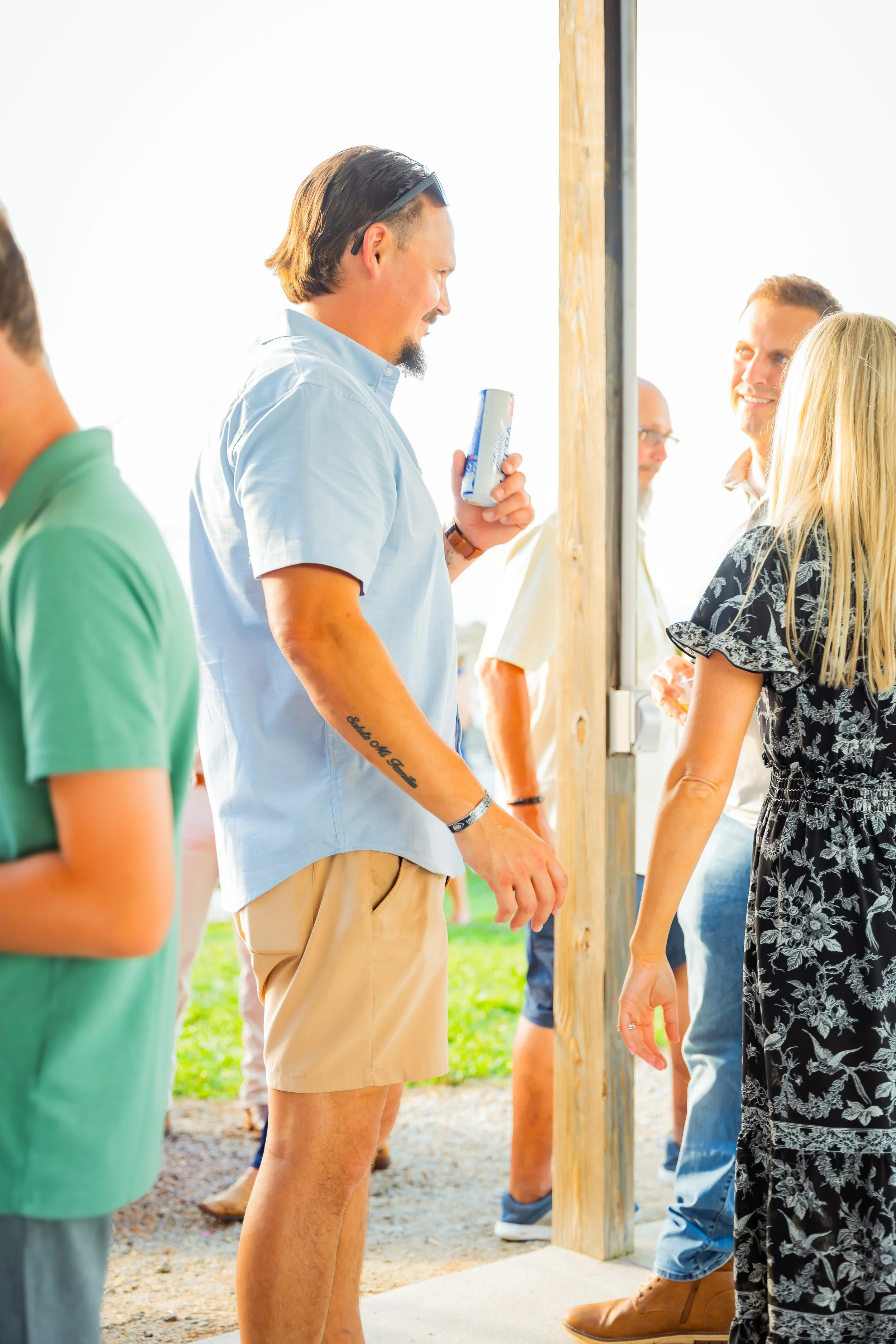 People shaking hands outdoors with a man holding a can, standing near a wooden post, smiling and engaging in conversation on a sunny day.