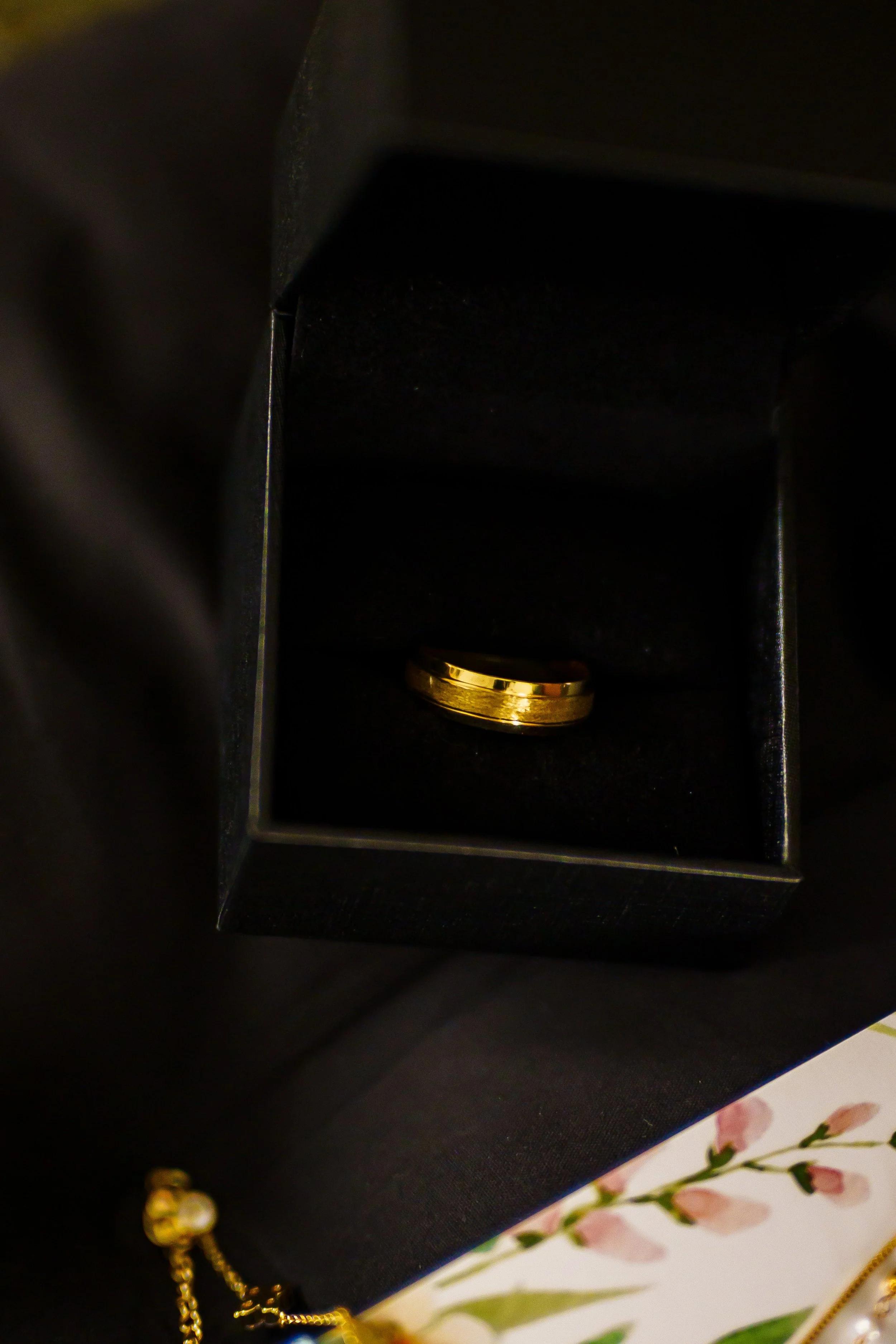 A black jewelry box with a gold ring inside and some jewelry and a floral patterned surface nearby.