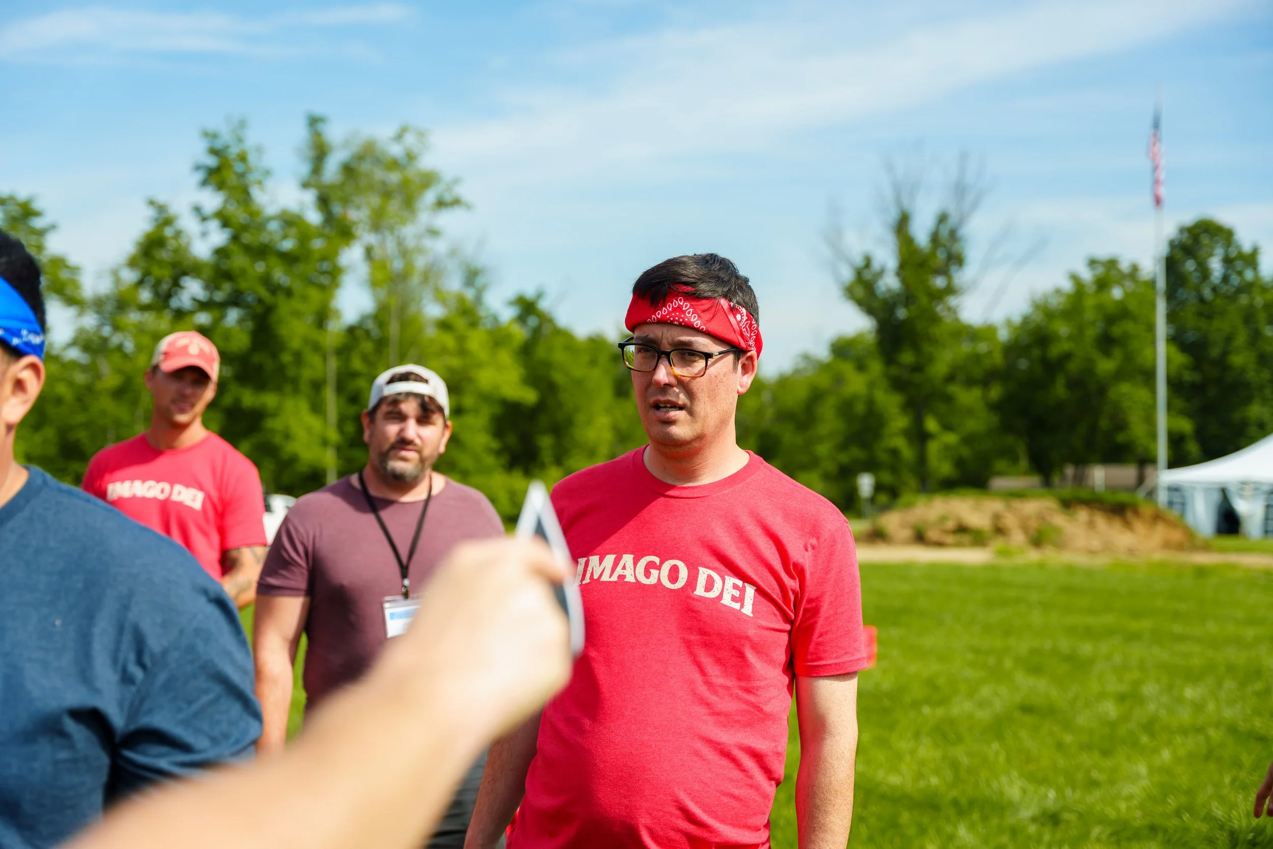 A group of men standing outdoors on a sunny day, one man in the foreground wearing glasses and a red bandana and red shirt with white text, others in the background wearing hats and casual clothing, surrounded by green trees and a partly cloudy sky.
