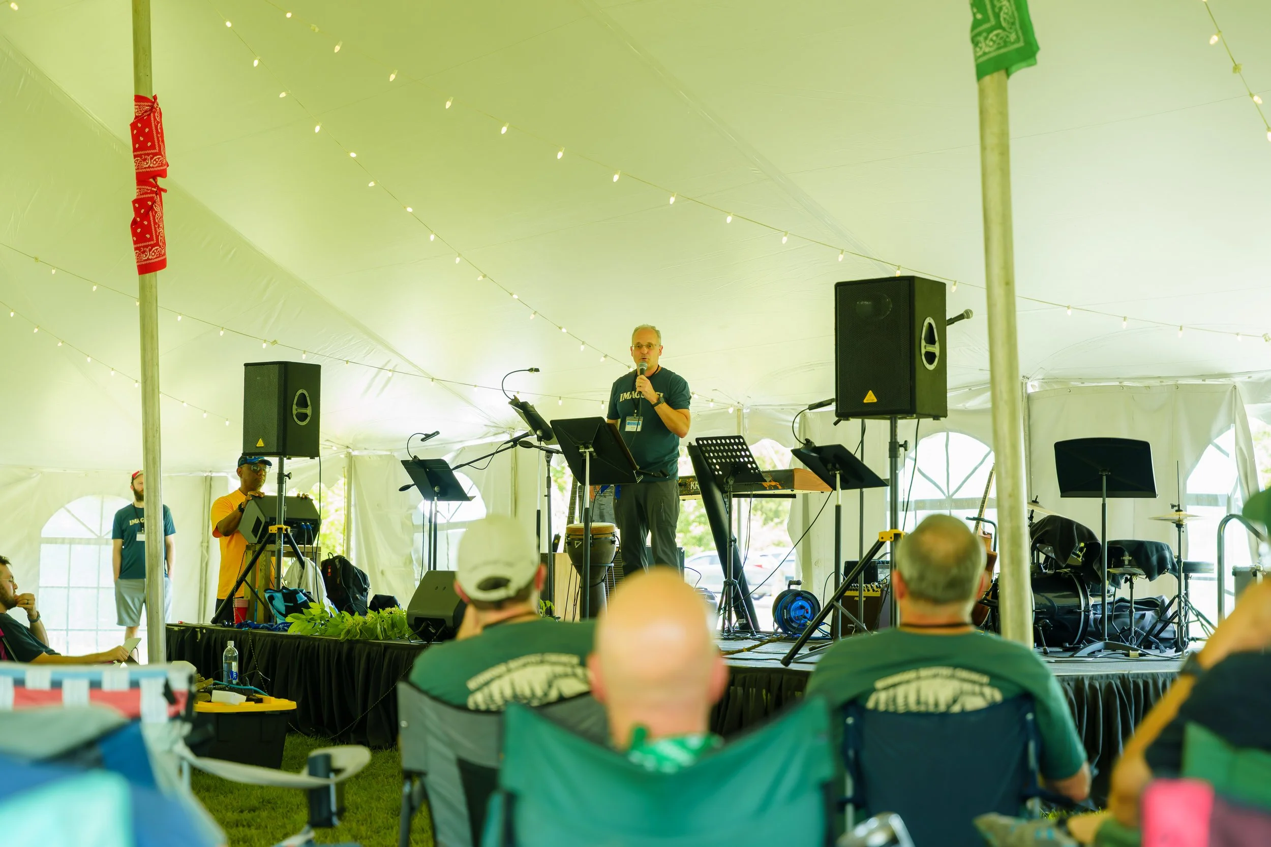 A speaker stands at a microphone on a stage inside a large green tent, addressing an audience. Musical instruments, speakers, and music stands are visible on stage. The audience is seated, and the tent is decorated with string lights and colorful fab