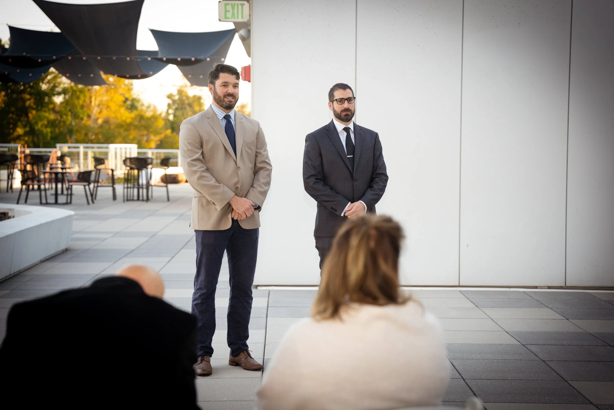 Two men stand in front of a white wall, dressed in business suits, with a group of people seated in front of them. The outdoor setting features trees with autumn foliage and tables with chairs in the background.