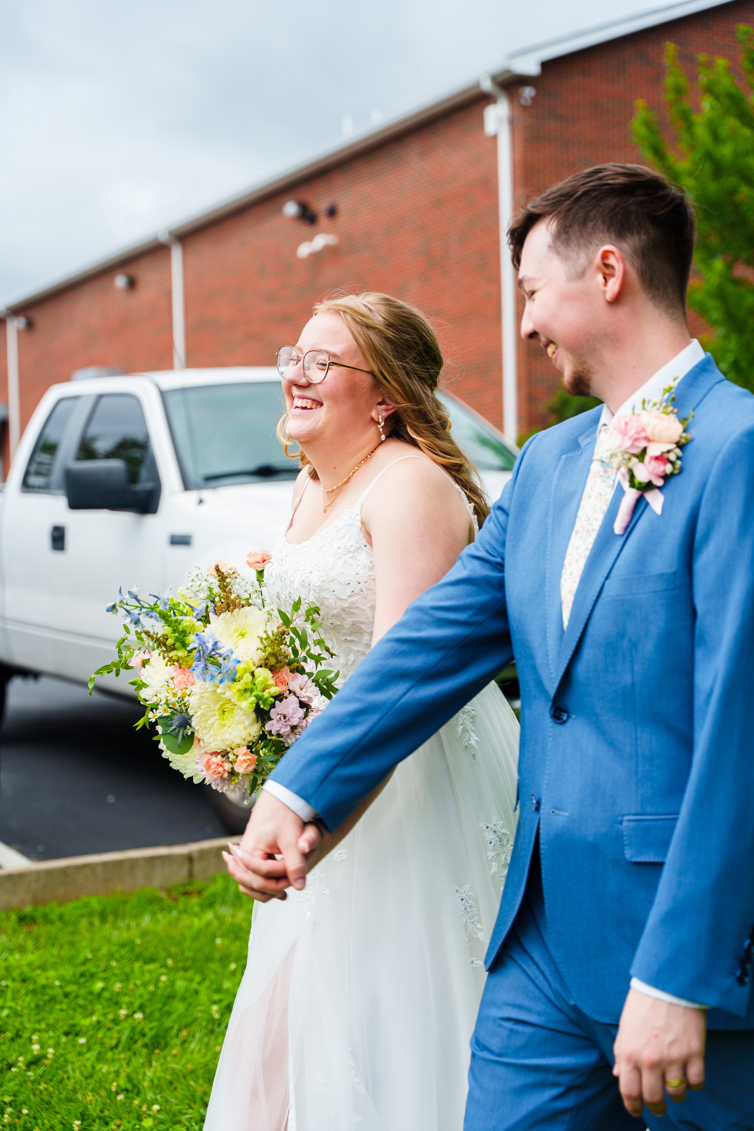 A bride and groom holding hands and smiling outside on a cloudy day, with a brick building and a white truck in the background. The bride is wearing glasses and a white dress, holding a large bouquet of colorful flowers. The groom is wearing a blue s