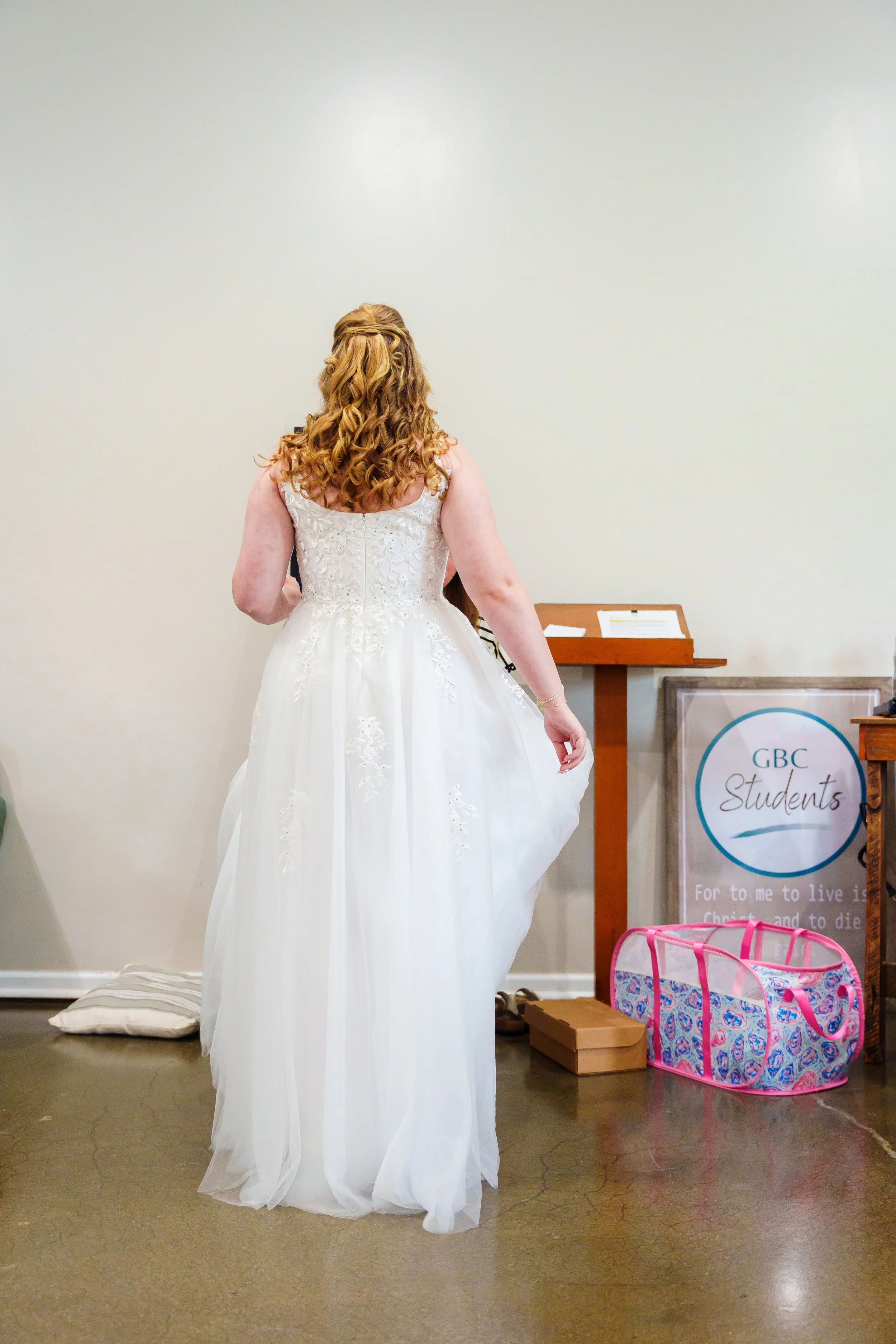 Back view of a woman wearing a white wedding dress with curly red hair in a half-up style, standing in front of a plain light-colored wall. There are various items on the floor and a sign on a stand that reads "GBC Students."