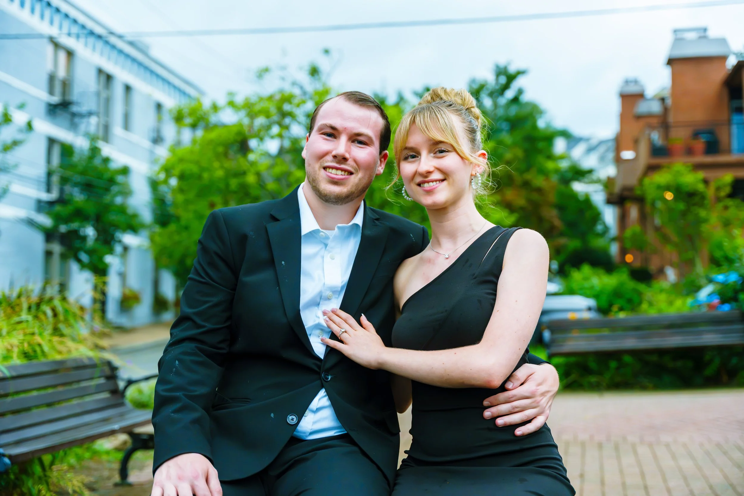 A smiling couple sitting on a park bench, dressed formally, with greenery and buildings in the background.