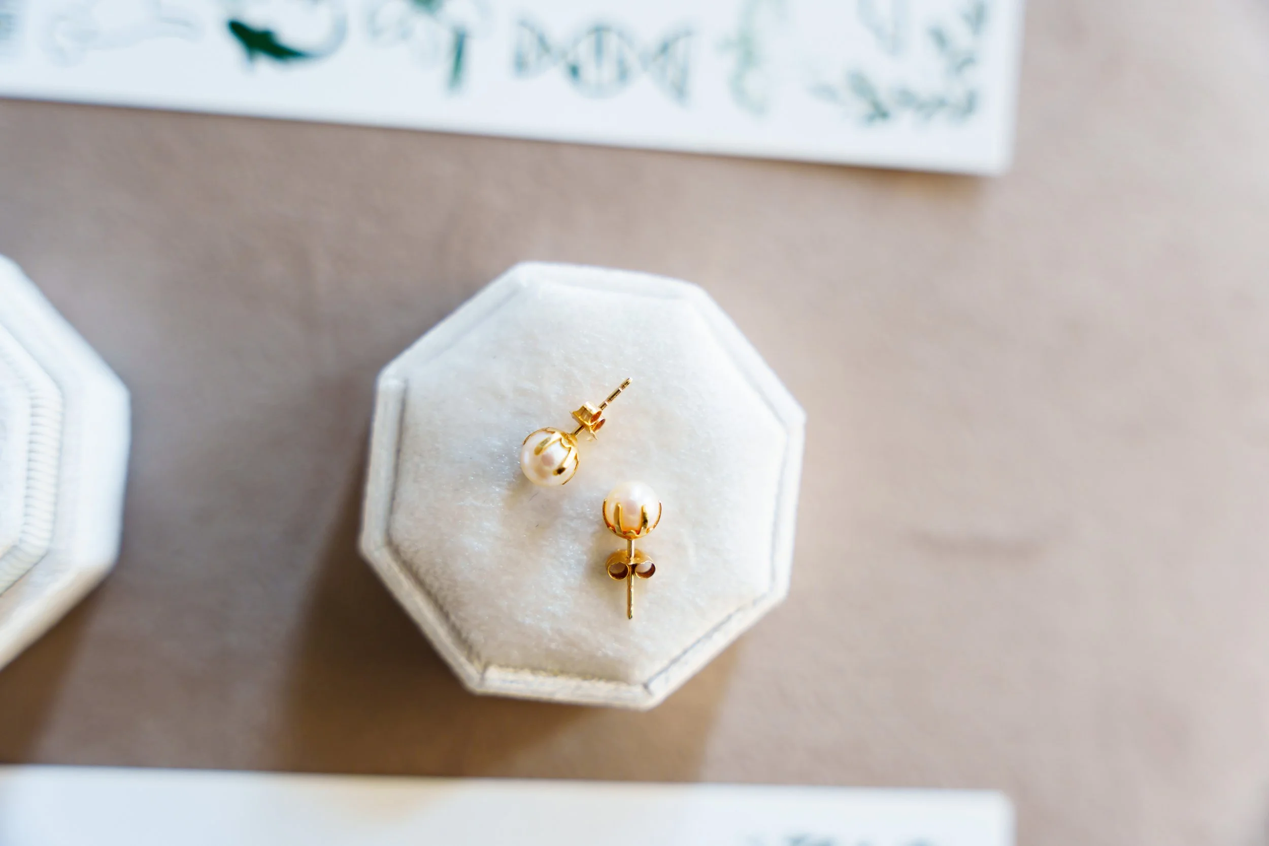 Gold and pearl earrings displayed on a white hexagonal jewelry box with a teal and white sign in the background.