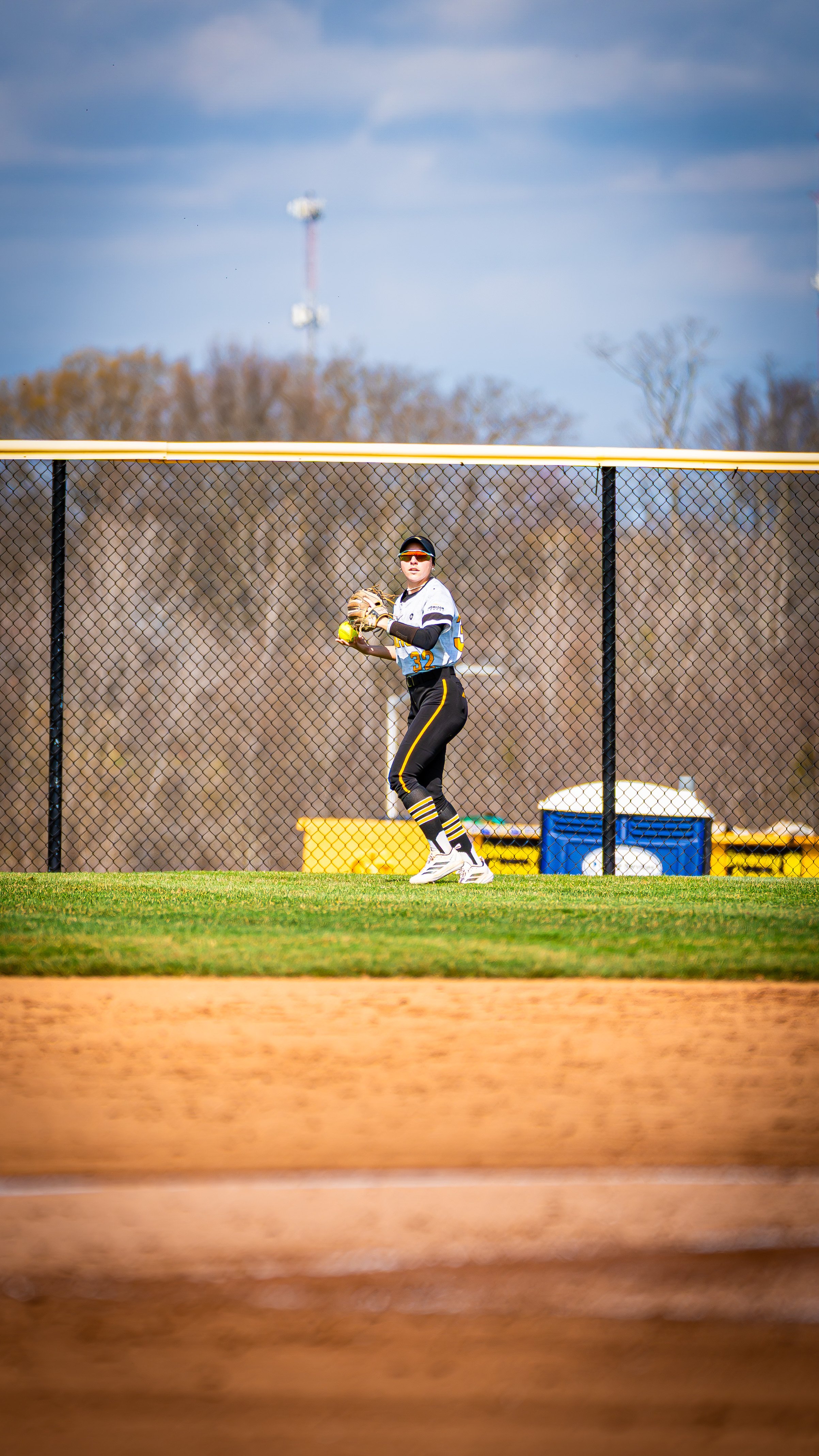 A young male baseball player in a white and black uniform with yellow accents standing on a baseball field, ready to catch a ball with a glove, behind a black chain-link fence with trees and utility poles in the background.