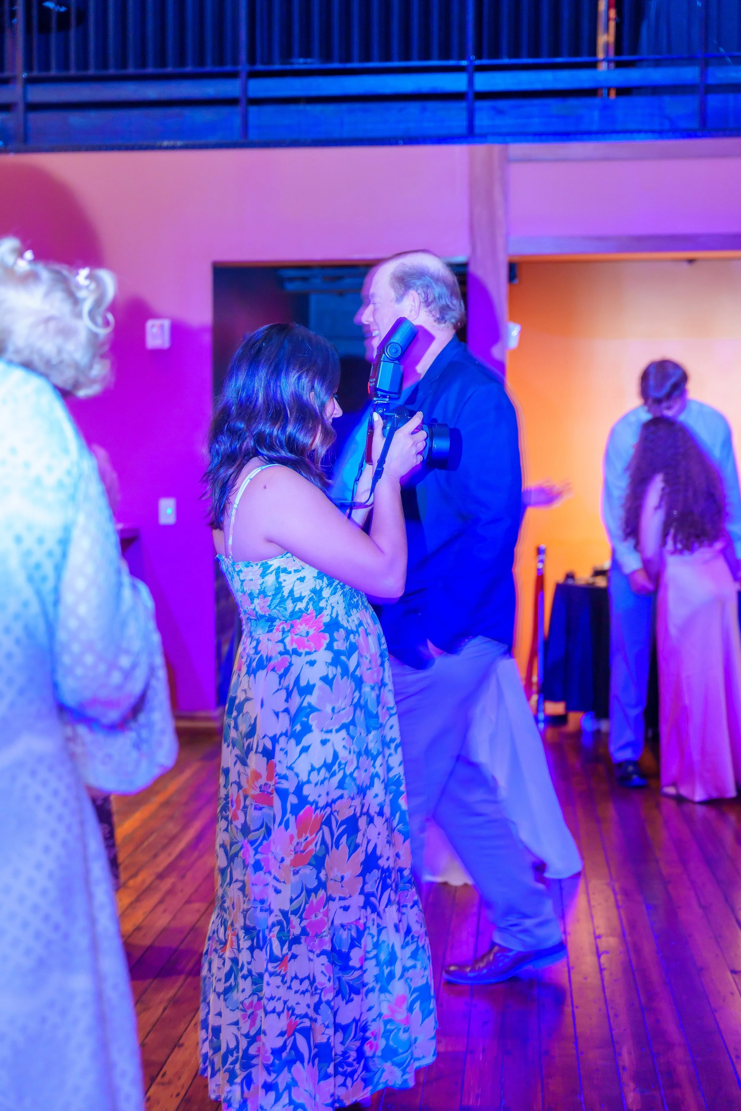A woman with shoulder-length wavy hair in a floral dress is taking a photo of an older man with gray hair in a dark blazer at a social event. Other people are in the background, with colorful lighting and a wooden dance floor.