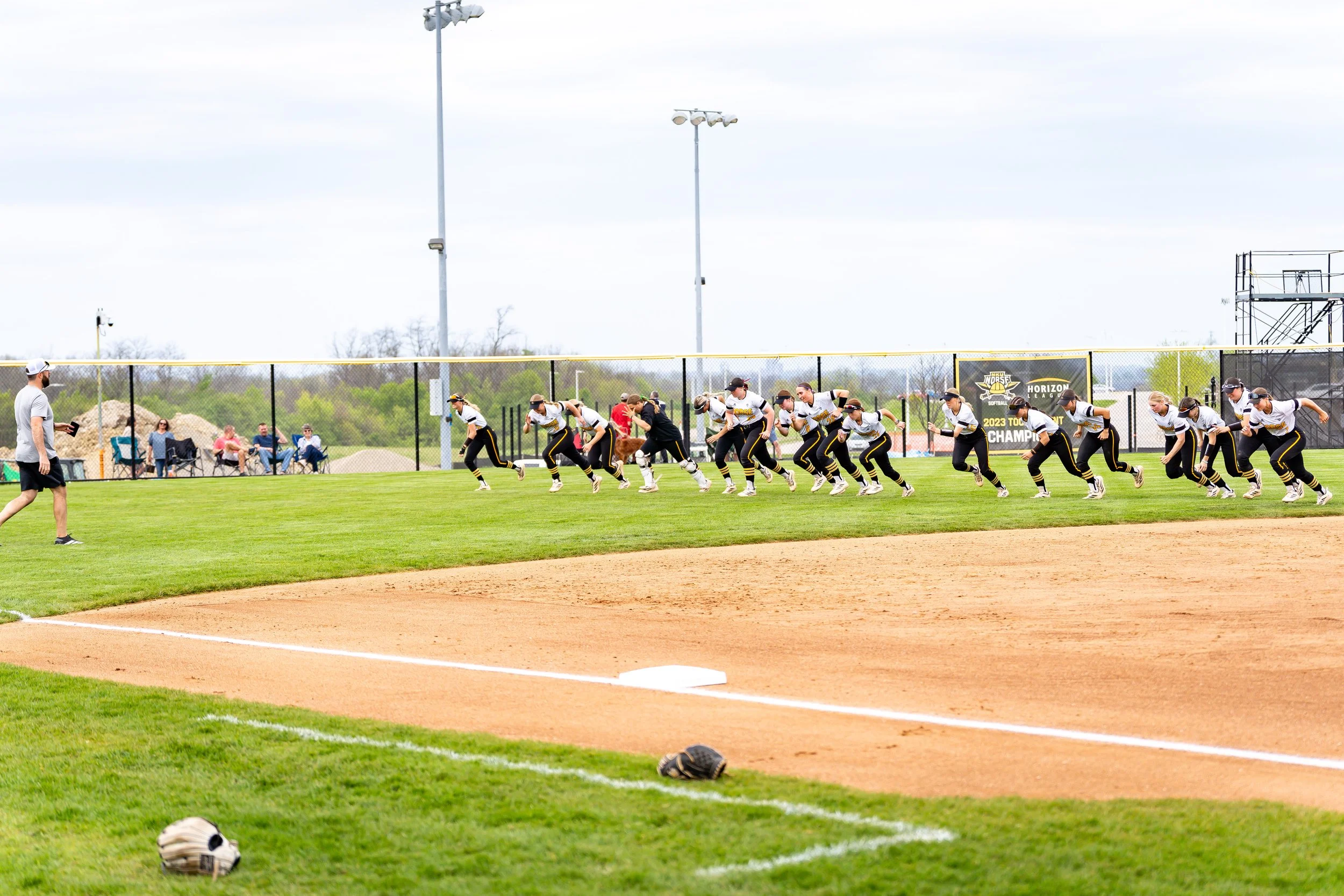 Softball team in white uniforms with yellow and black accents huddles on the field during a game. A coach stands nearby with a clipboard, and behind them is a fence with a banner that reads "2023 Tournament Champions." Spectators are seated on benche