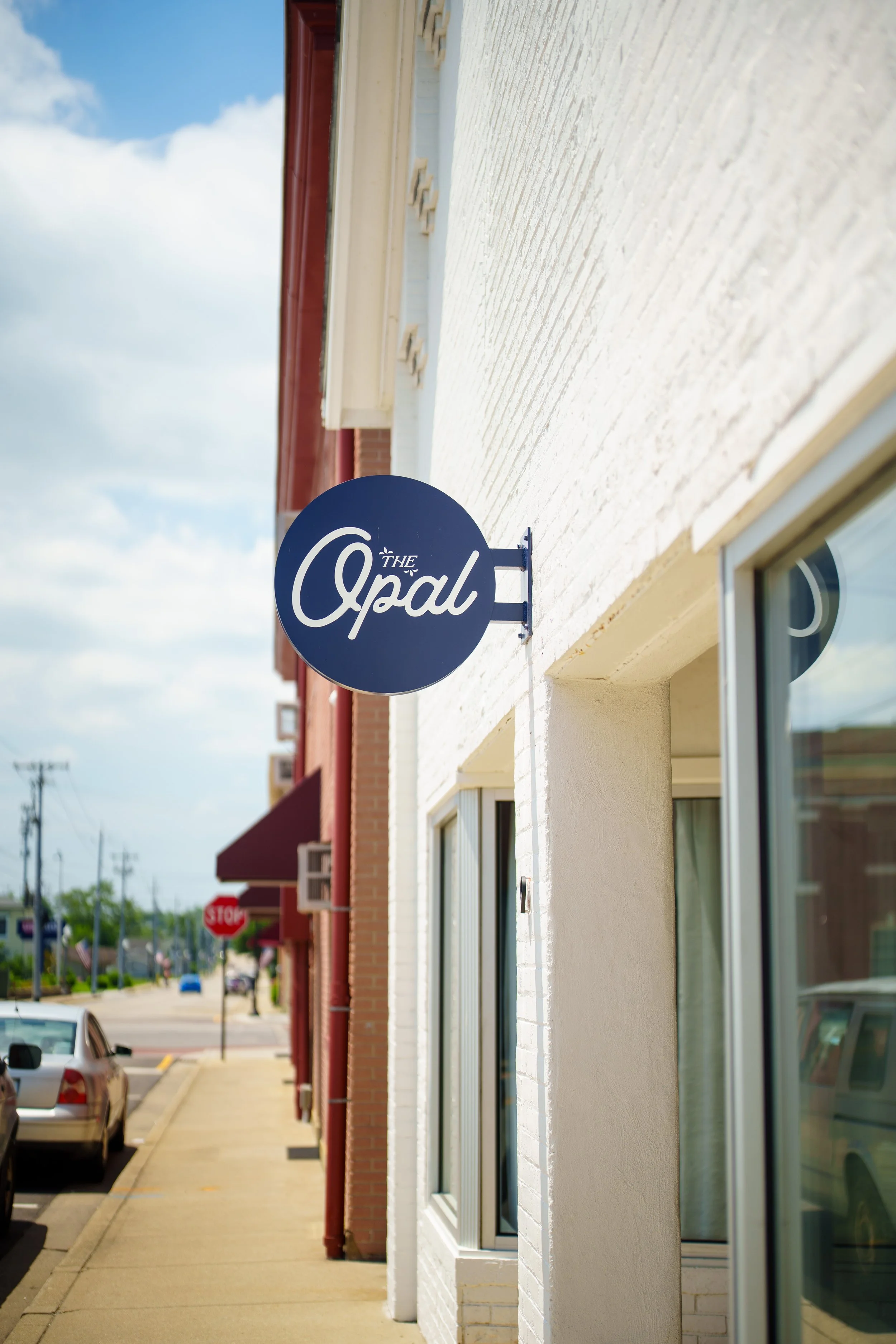 A sidewalk with parked cars along a white brick building featuring a circular blue sign with white text that reads 'The Opal'. The sky is partly cloudy, and there is a red brick building with a burgundy awning further down the street.