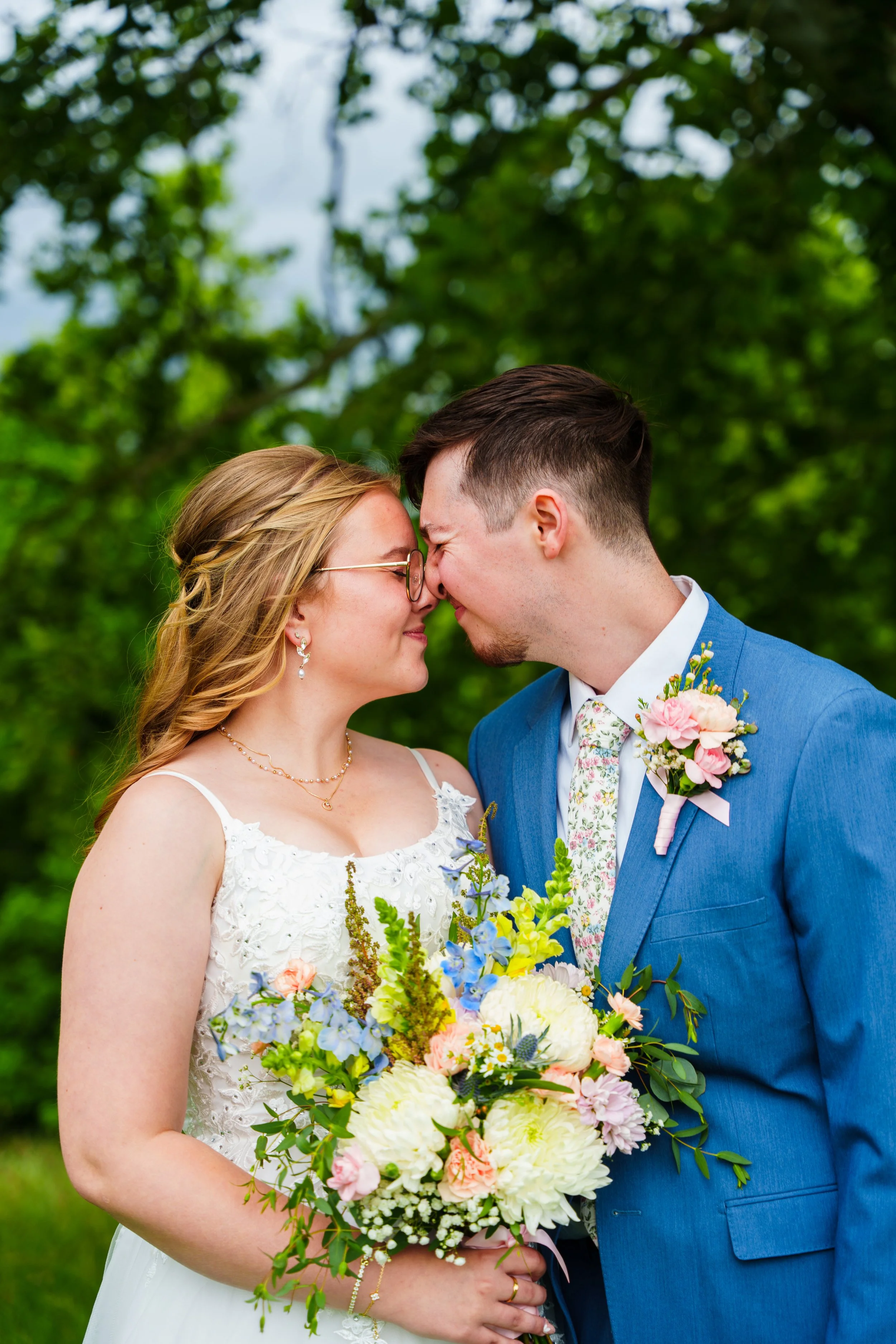 A bride and groom touching foreheads during a wedding outdoors, with greenery in the background. The bride wears glasses and a white lace dress, holding a colorful bouquet. The groom wears a blue suit with a floral tie and boutonniere.