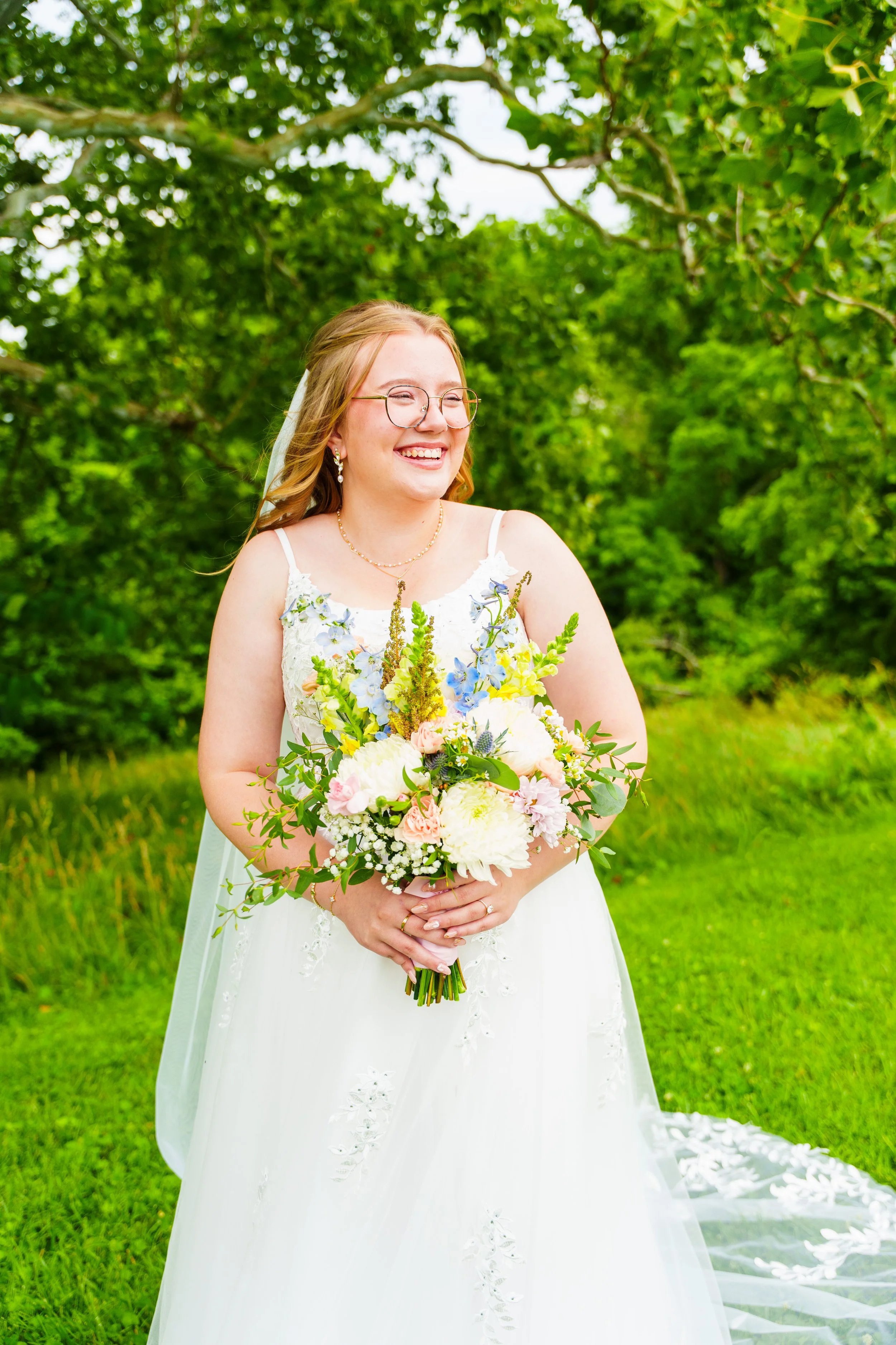 A bride with long blonde hair, glasses, and earrings standing outdoors with green trees behind her, holding a large colorful bouquet of flowers, smiling.