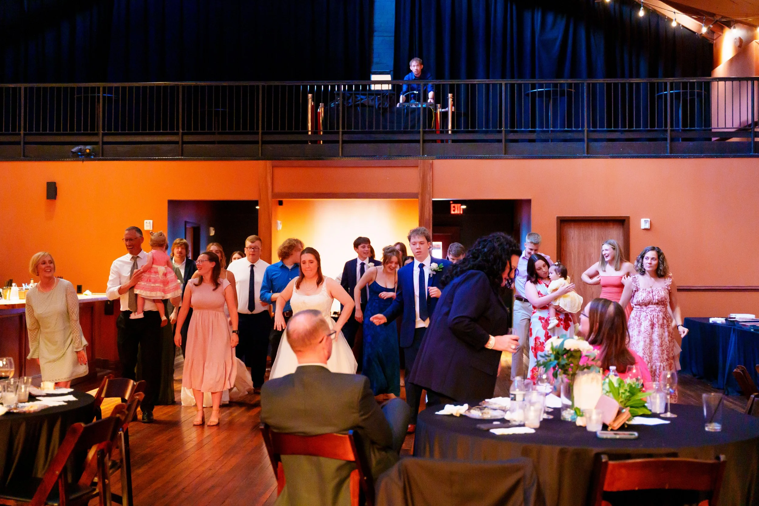 People attending a formal event in a decorated venue with tables and a stage, some are standing and mingling, with a DJ or speaker on stage and string lights hanging from the ceiling.