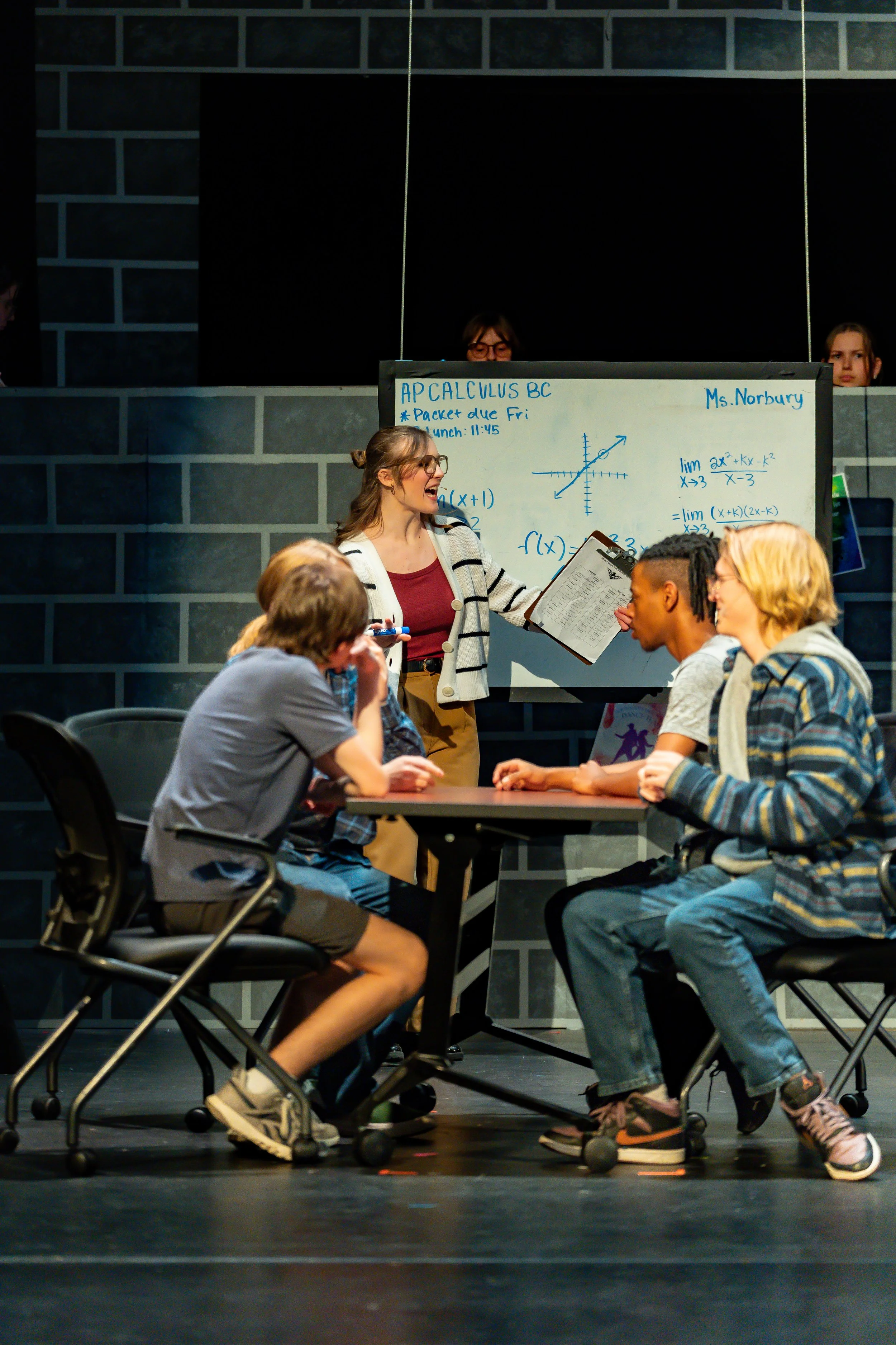 A classroom scene showing a young woman teacher standing near a whiteboard with math notes, talking to students seated at a table, with two students visible in the foreground and two more students partially behind the whiteboard. The background featu