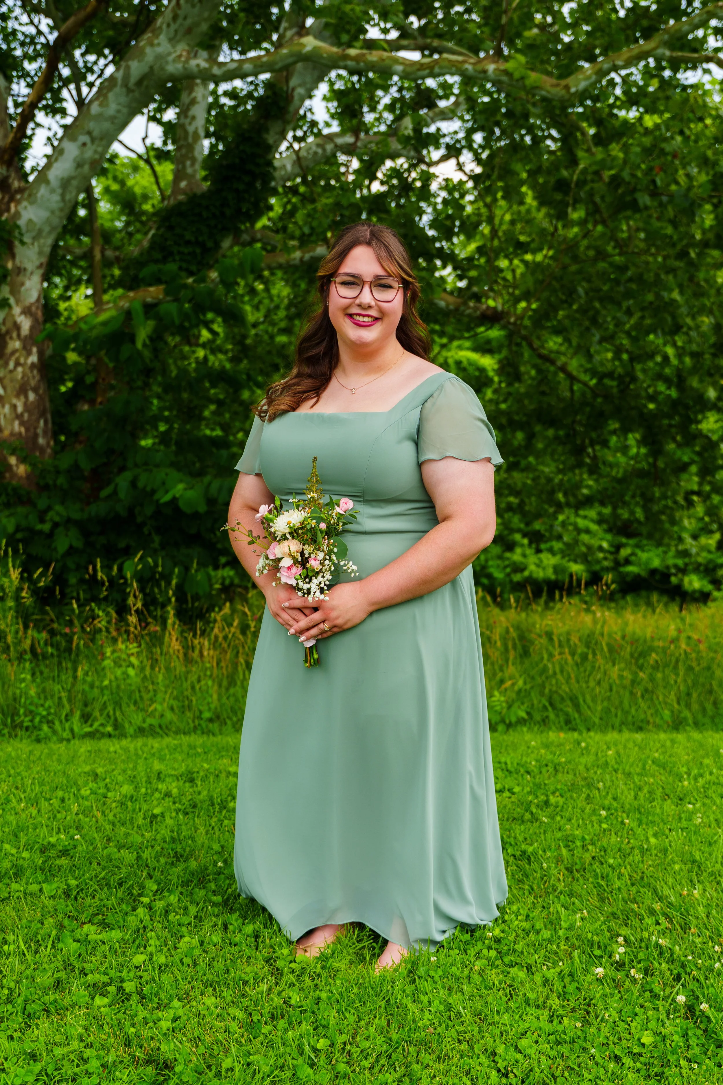 A young woman with glasses in a green dress holding a bouquet of flowers standing on grass with lush green trees in the background.