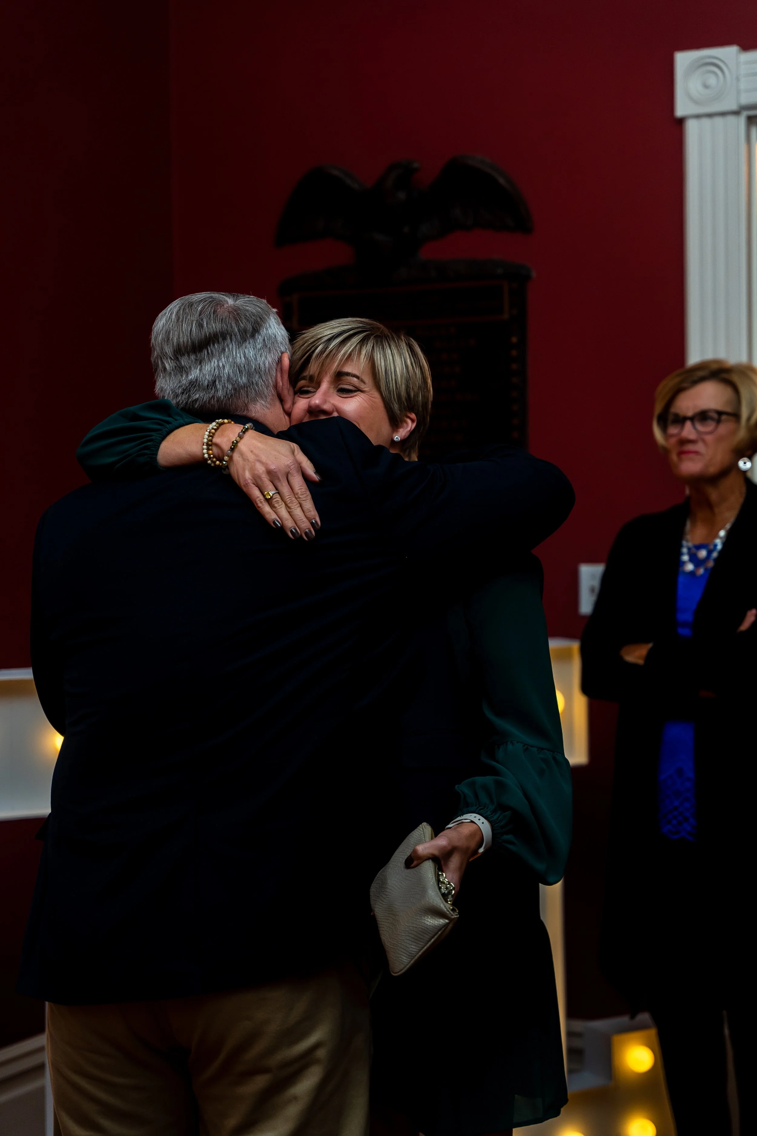 A woman hugging an older man in a dark room with red walls, another woman with glasses watching in the background.