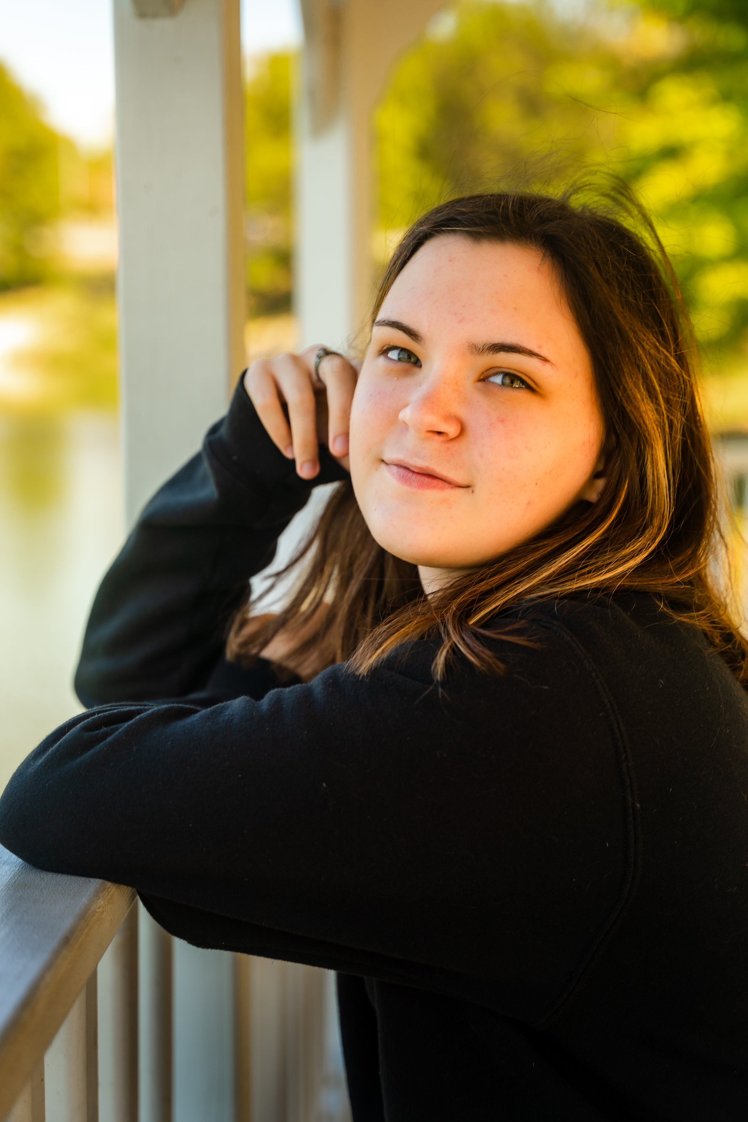 Close-up of a young woman with light skin and brown hair, smiling slightly, wearing a black sweatshirt, standing outdoors near a railing with a blurred background of green trees and yellow foliage.