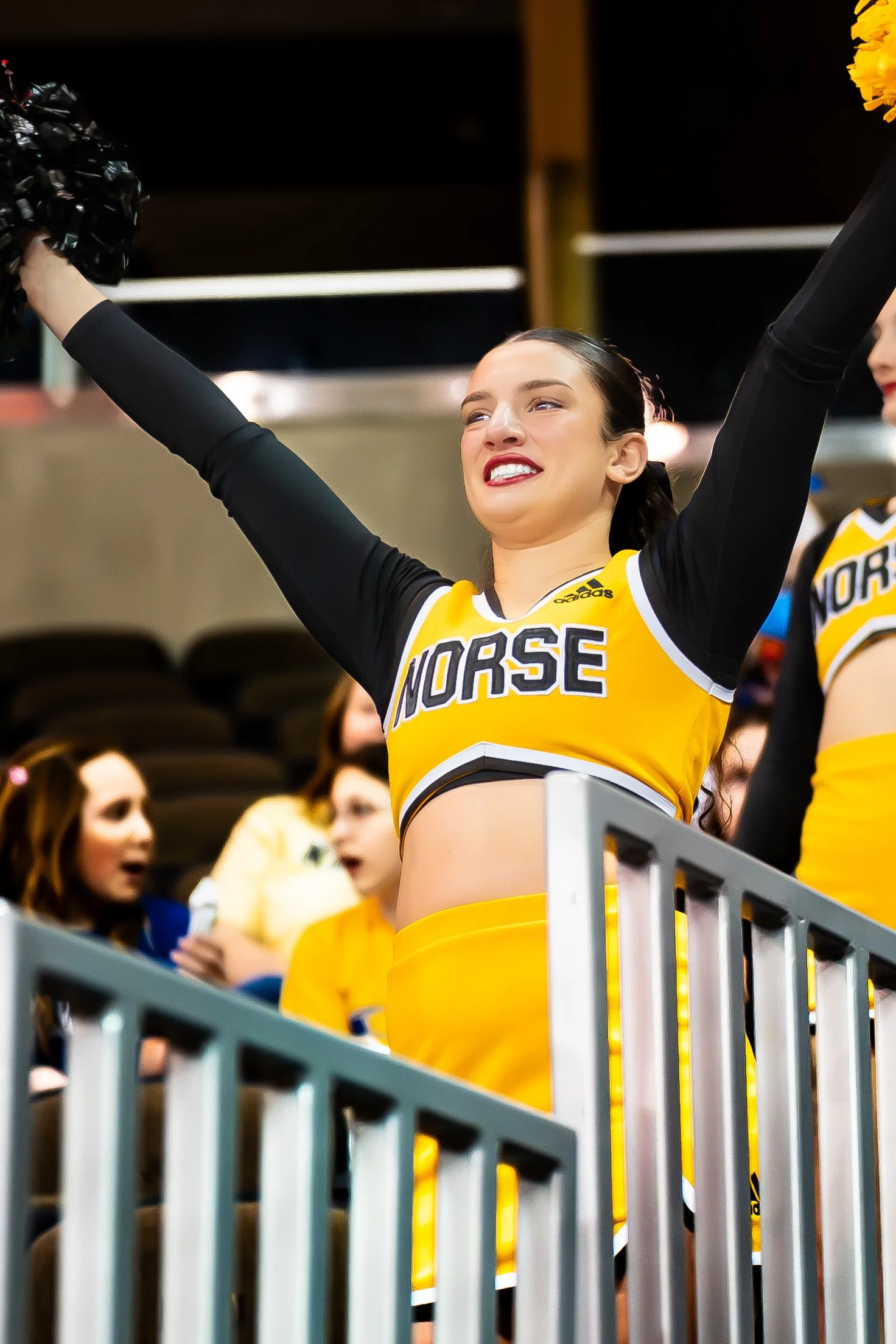 Cheerleader in yellow and black uniform with 'Norse' written on it, raising arms in celebration at a sports event.