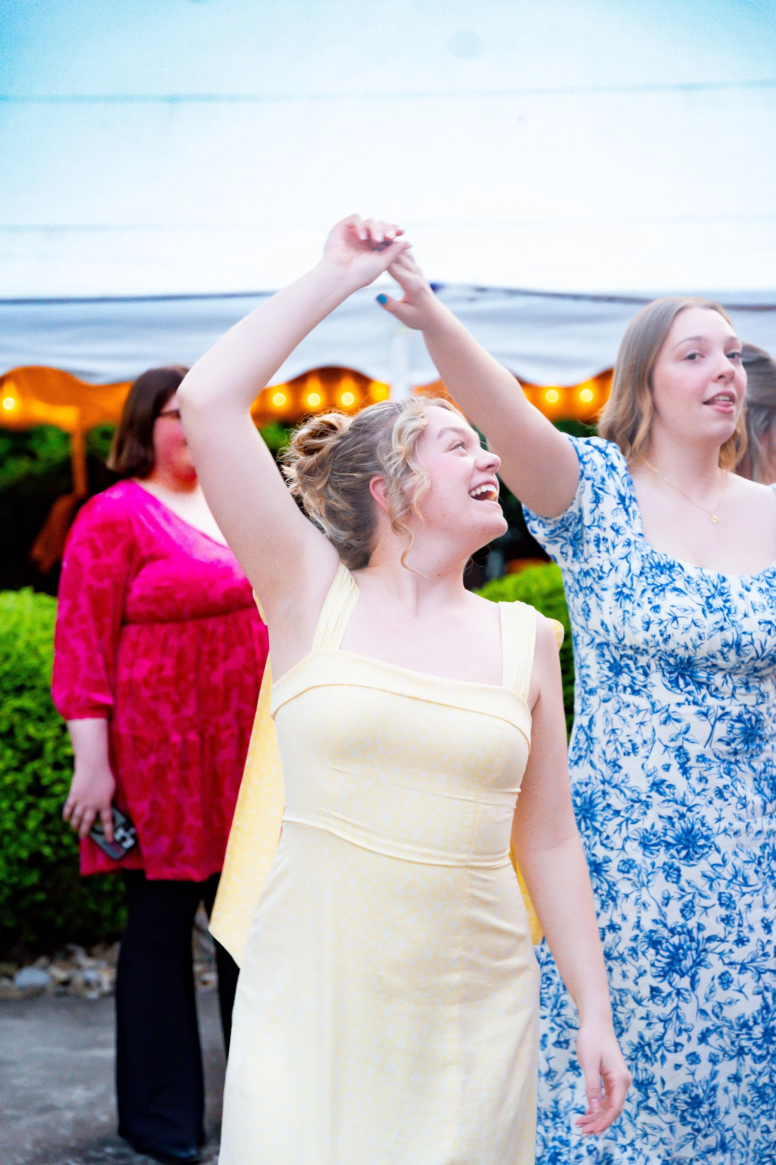 Women dancing at an outdoor event with a white tent and string lights in the background.