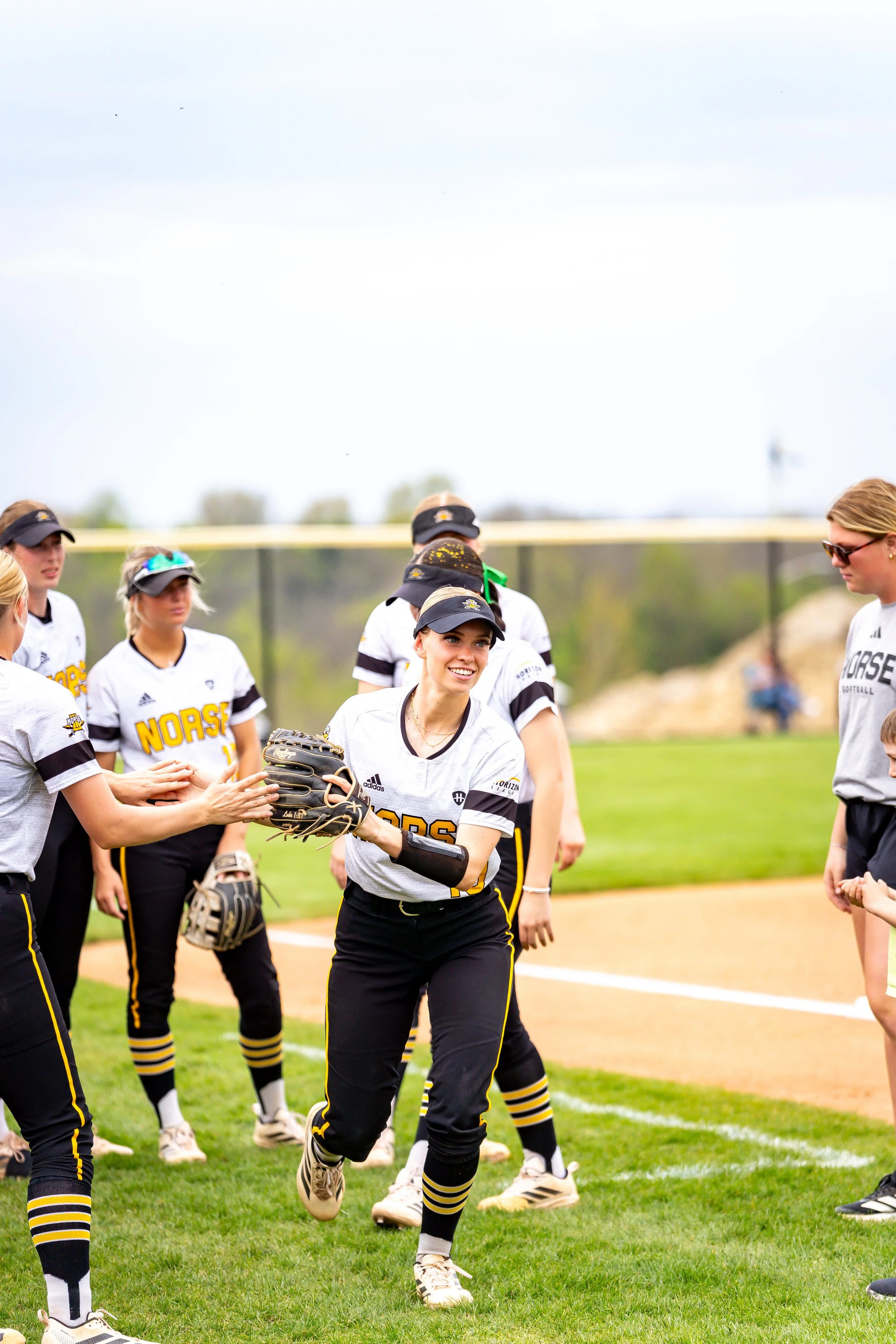 Softball players on a field, wearing white jerseys with black and yellow accents, gathering before or during a game, with a cloudy sky and trees in the background.