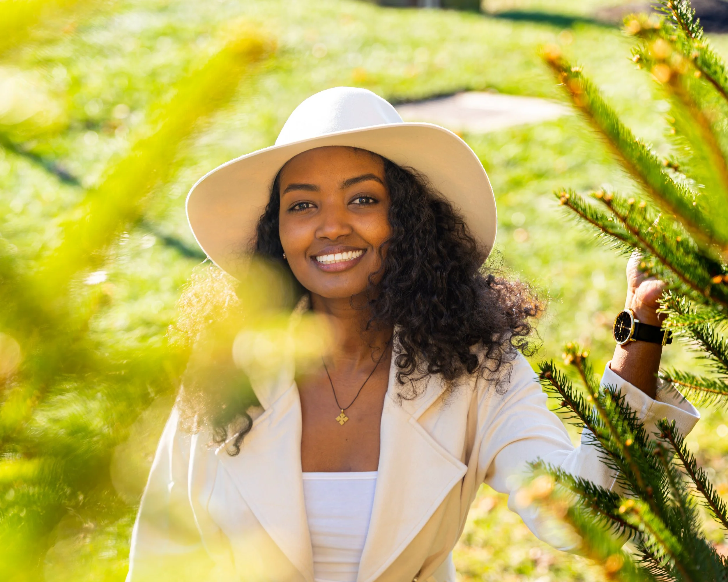 Smiling woman wearing a white hat and white jacket outdoors, surrounded by greenery and pine branches.