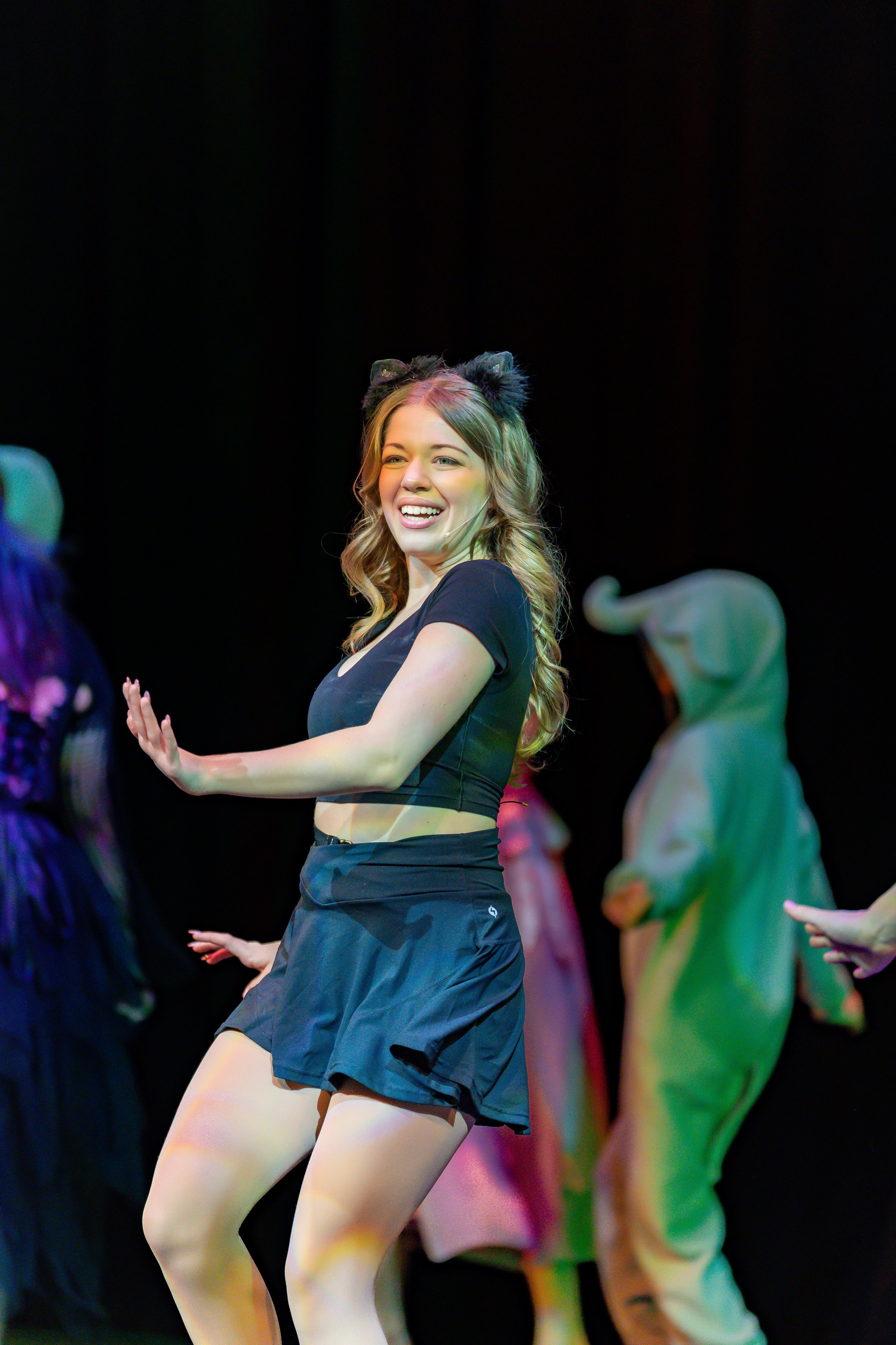 A young woman with wavy blonde hair, wearing a black crop top and black shorts, performing a dance move with her arms outstretched. She is on stage with several other dancers in colorful costumes in the background, under stage lighting.