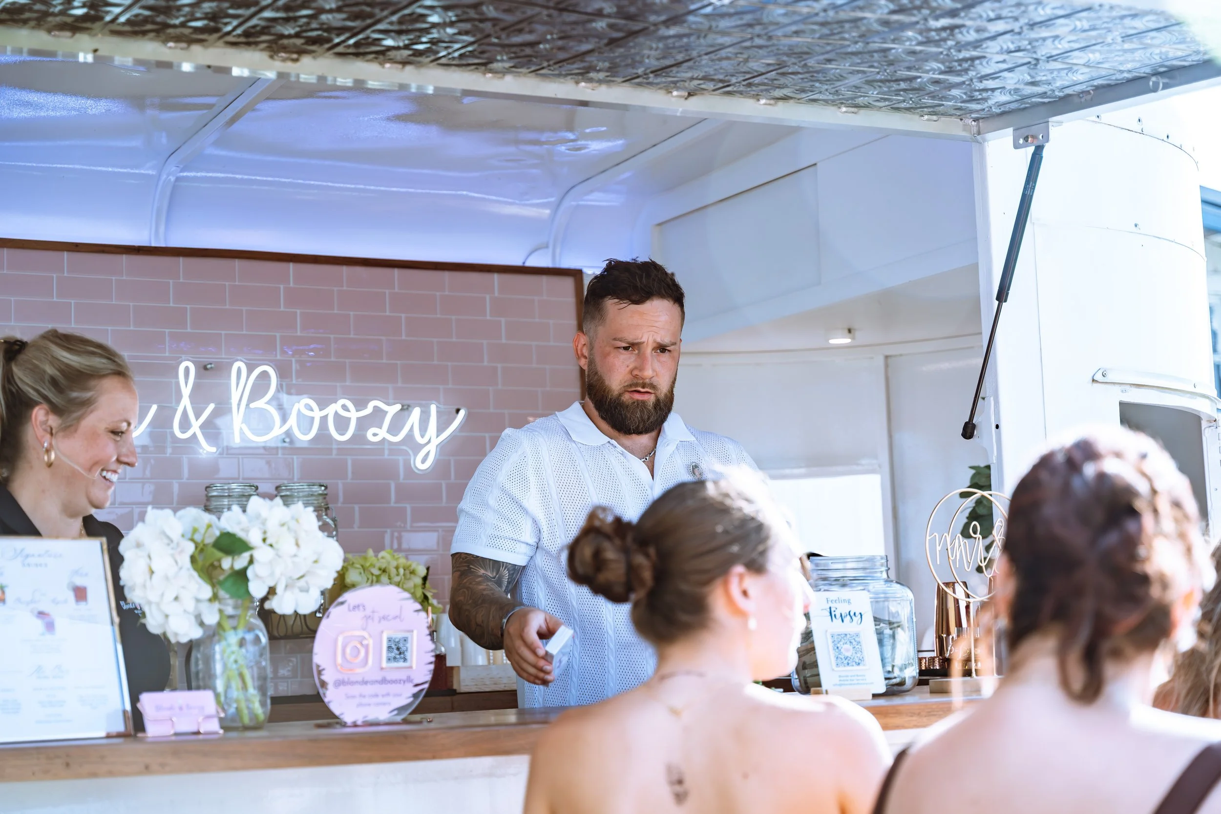 A man with a beard and tattoos wearing a white shirt working at a wedding reception food truck. The background features a pink brick wall and a neon sign that reads 'Eat & Boozy.' There are flowers, menus, and QR codes on the counter, with guests in 