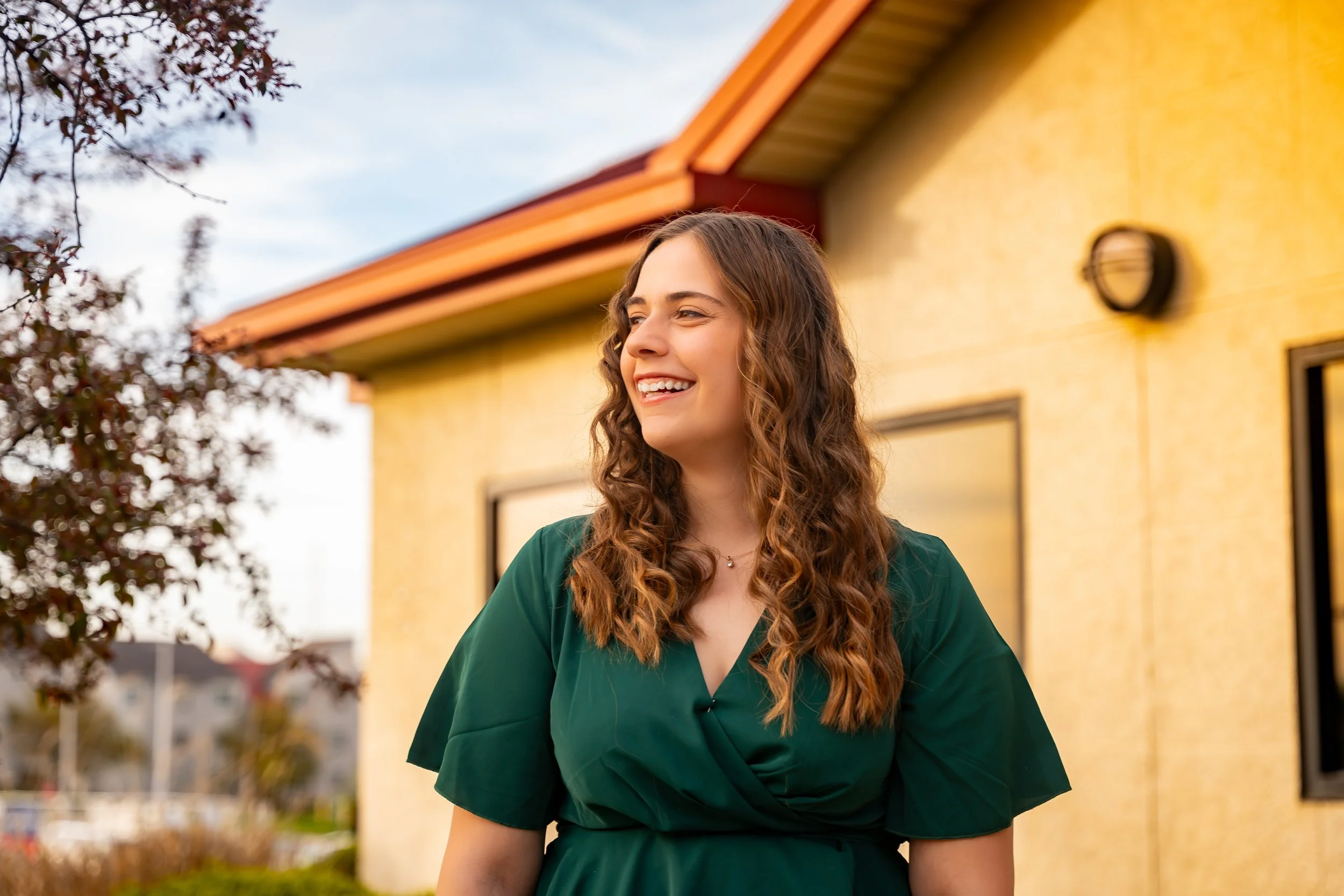 A young woman with long, curly brown hair smiling and wearing a dark green dress, standing outside near a yellow house with a red roof, with a tree with dark leaves in the foreground and a cloudy sky in the background.