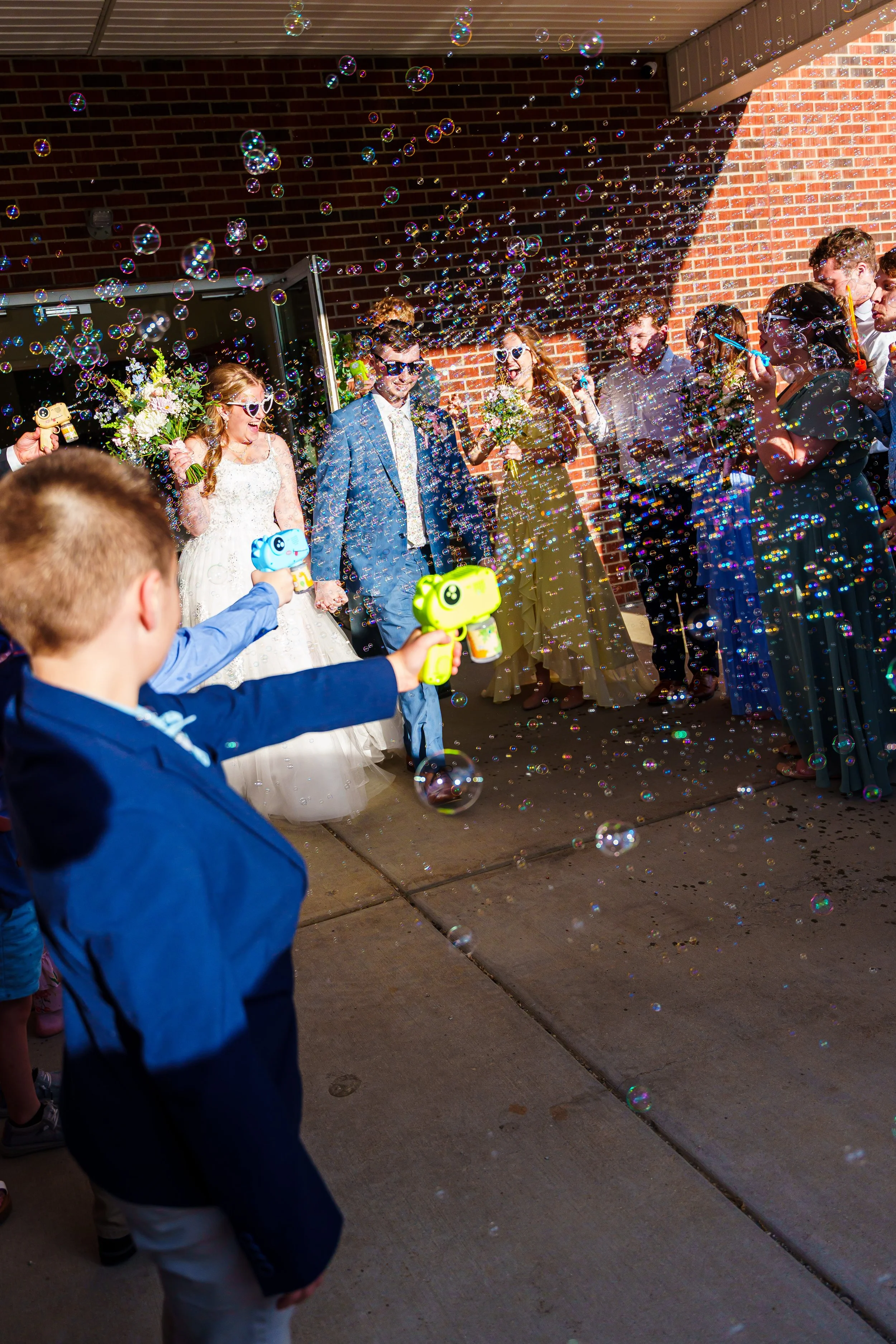 People at a party celebrating with bubbles, including a bride in a wedding dress, a groom in a suit, and guests wearing sunglasses, holding water guns, and laughing against a brick wall.