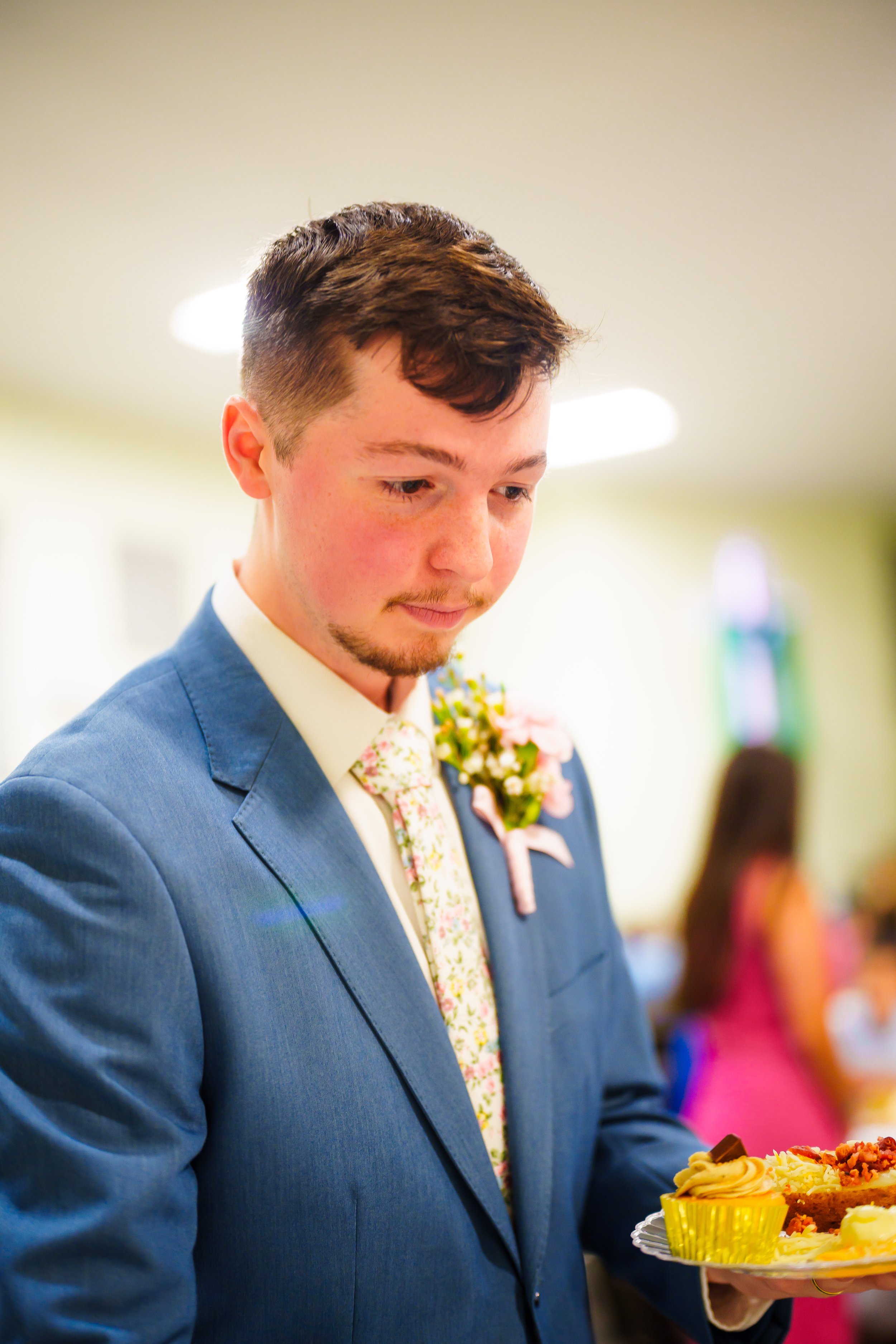 A young man in a blue suit is holding a plate of assorted desserts at a social event, with a blurred background of other people.