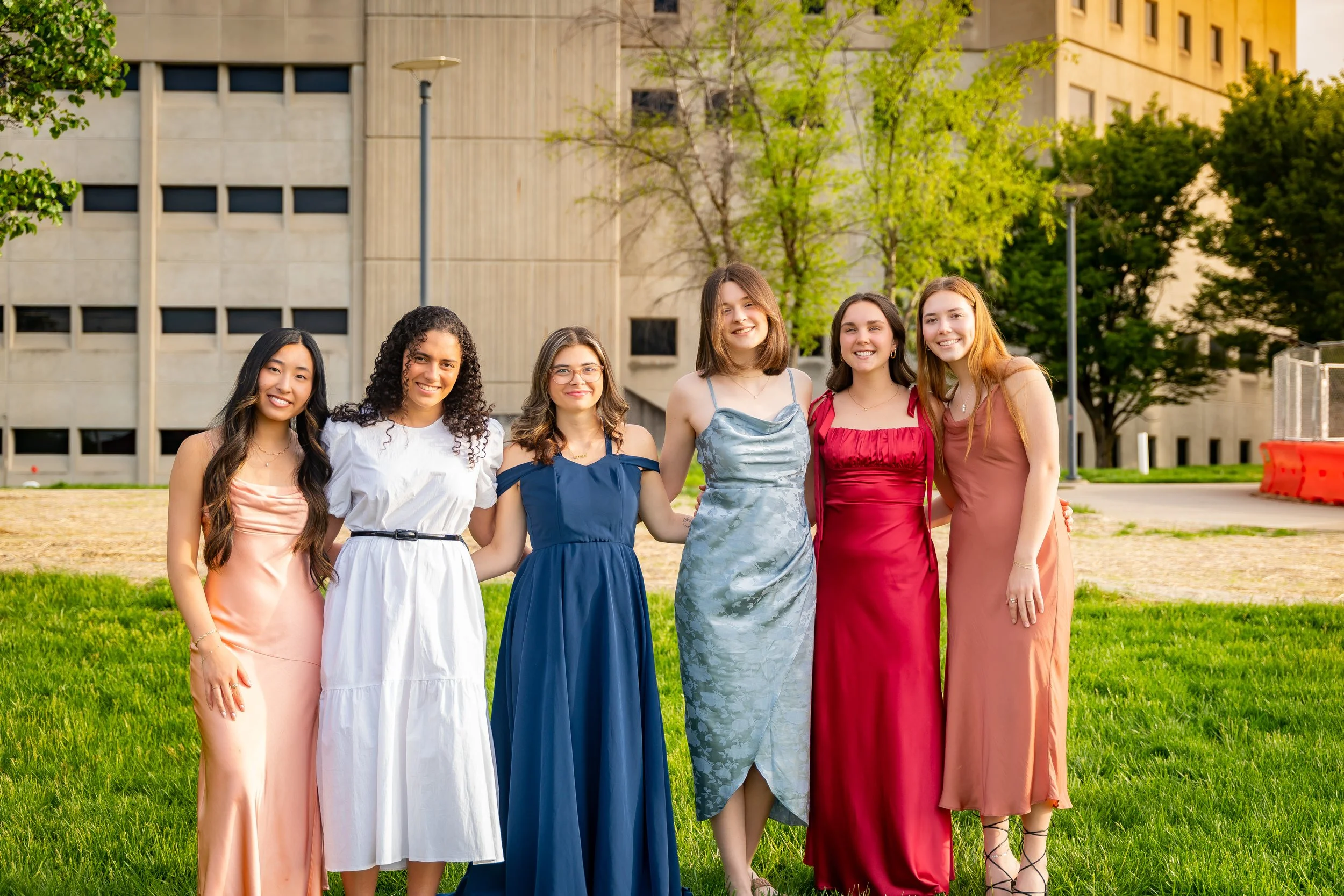 A diverse group of six young women in colorful dresses standing outdoors on a grassy area, smiling, with trees and a building in the background.