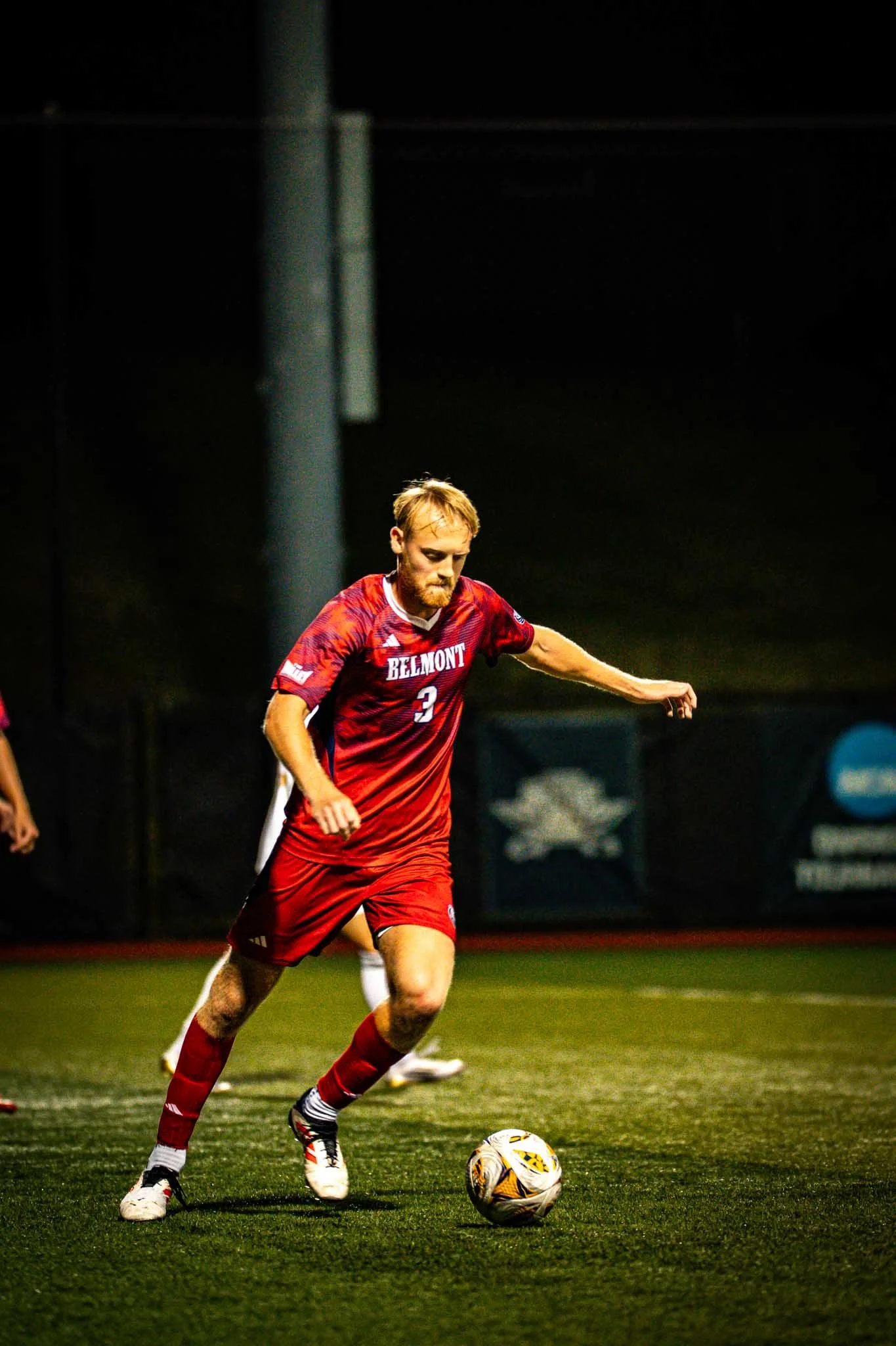 A male soccer player with a beard in a red Belmont jersey is dribbling a soccer ball on a field at night, with other players partially visible around him.