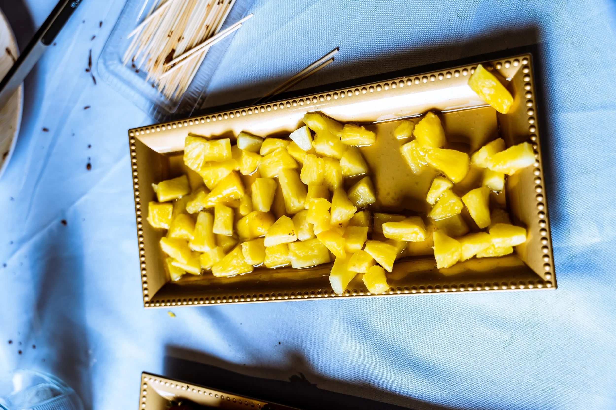 A rectangular metal tray filled with chopped yellow pineapple pieces on a light blue tablecloth, with a small portion of an empty tray visible in the top left corner.