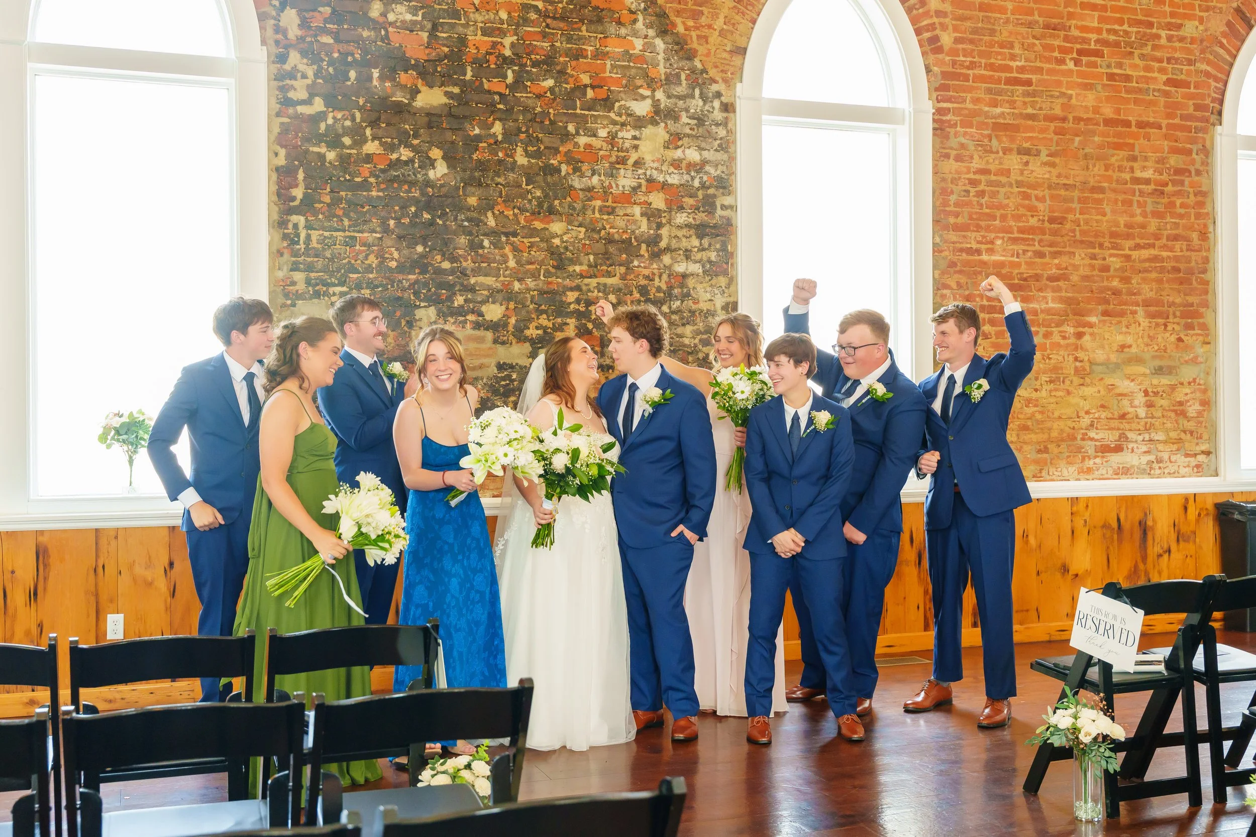 A group of people dressed in formal attire, celebrating at a wedding ceremony in a room with large arched windows, a brick wall, and wooden accents.
