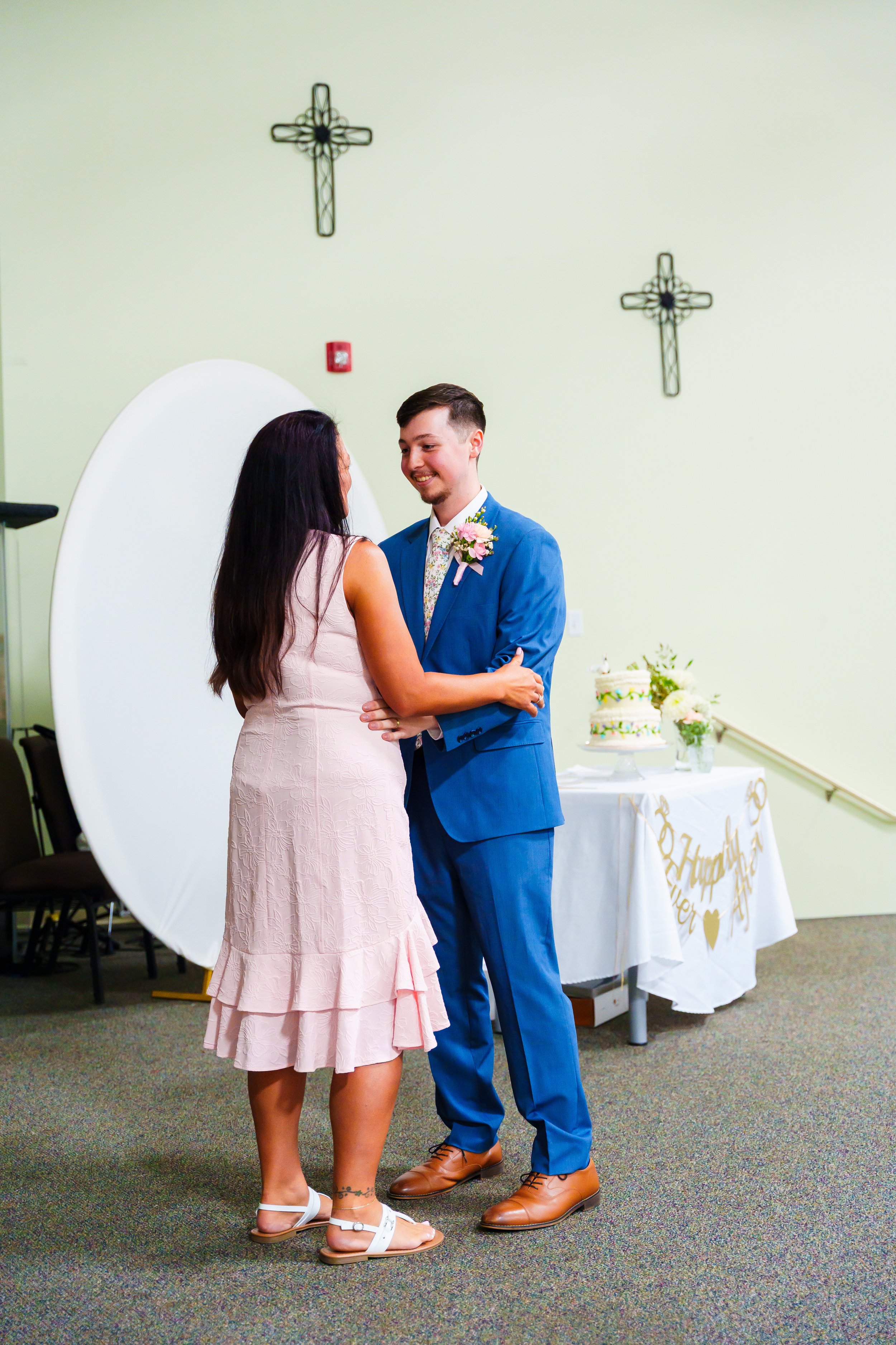 A couple celebrating a wedding or special occasion indoors, with the woman wearing a pink dress and the man in a blue suit, holding hands and smiling at each other. In the background, there is a decorated table with a cake and flowers, and a sign wit