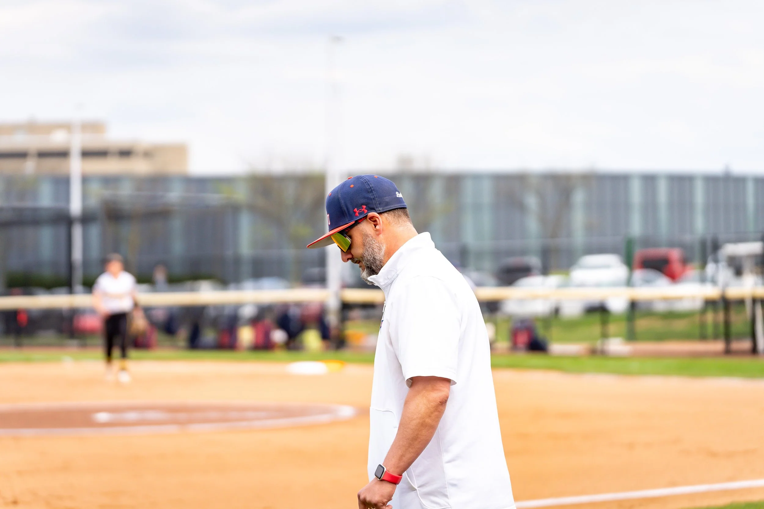 A man with a gray beard wearing a blue cap with red accents and sunglasses, dressed in a white shirt, stands on a baseball field with a fence, other people, and a cityscape in the background.