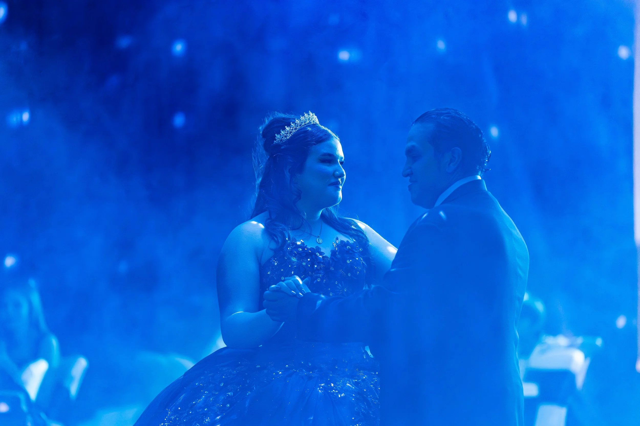 A couple dancing closely during a wedding, illuminated by blue lighting, with the bride wearing a tiara and a diamond necklace, and the groom in a suit.