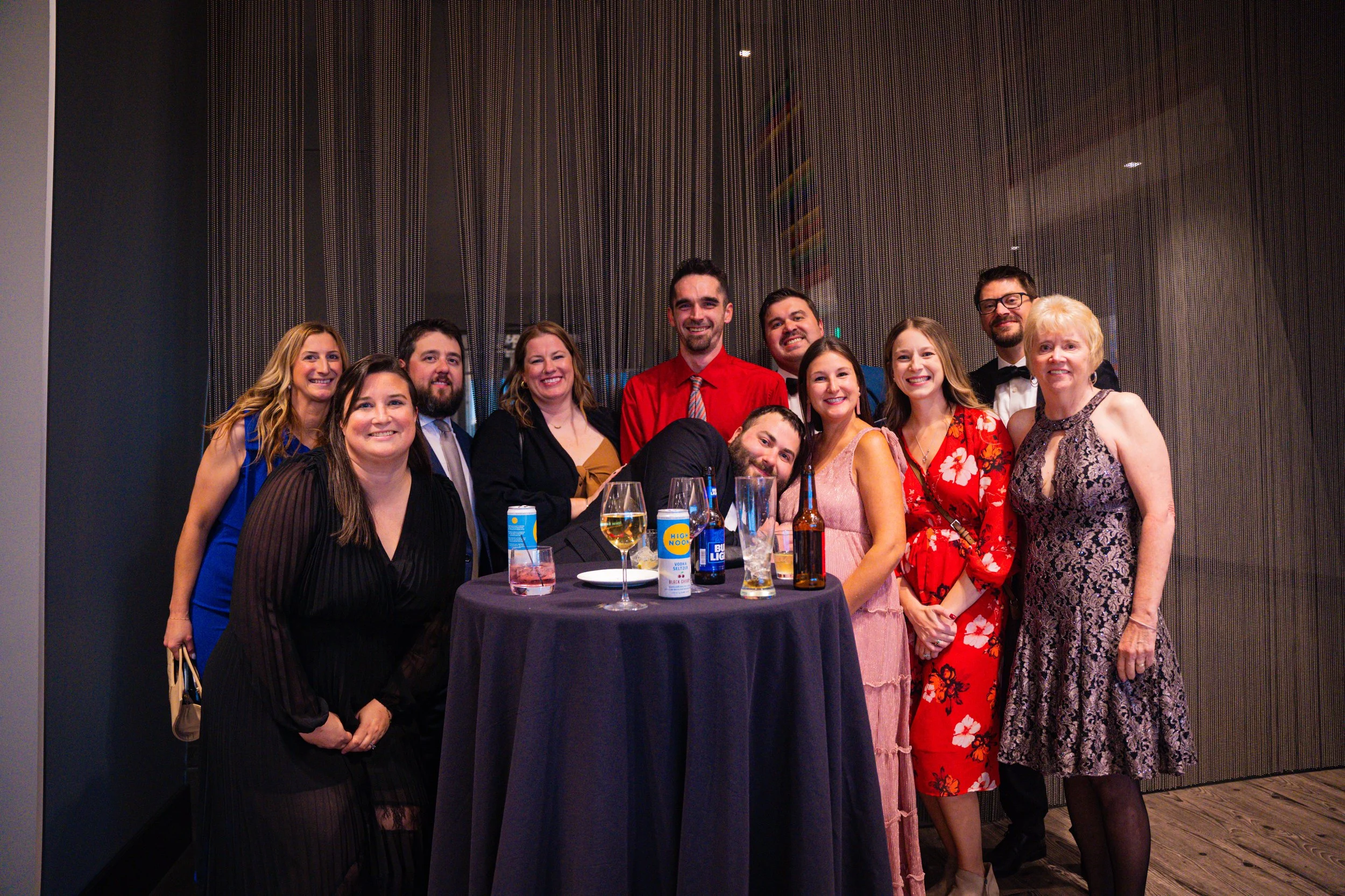 A group of eleven people, including men and women, dressed formally and smiling, gathered around a table at an indoor event. The table has drinks including wine and beer, and the background features dark curtains or wall paneling.